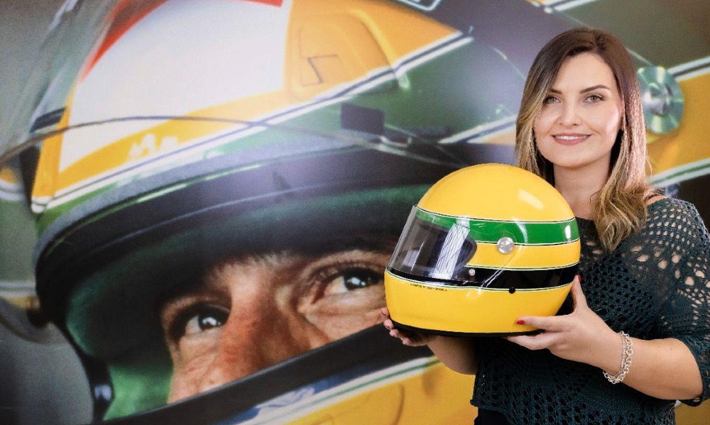 Uma mulher loira está segurando o capacete verde e amarelo de Ayrton Sena e posando para a foto, à frente de uma parede com uma foto enorme do ídolo nacional usando o mesmo capacete