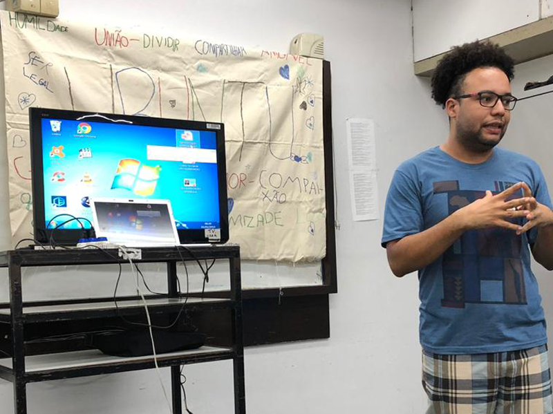 Um professor está em pé falando para alunos (que não vemos na imagem) e ao fundo está o quadro branco e um televisor conectado a um computador