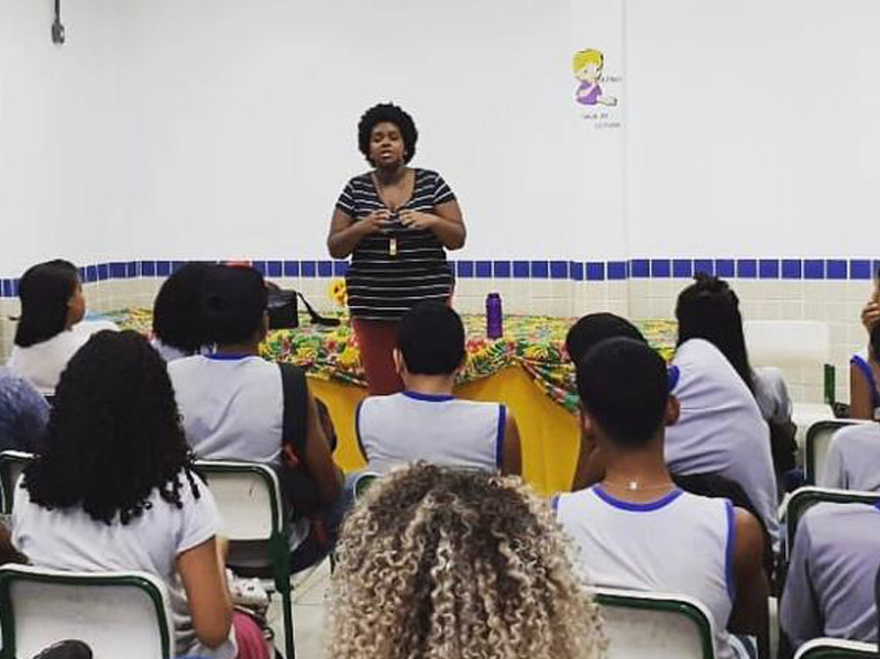 Imagem feita de uma sala de aula cheia de crianças sentadas e de costas para quem fez a foto. Elas estão ouvindo a professora, que está em pé à frente, falando algo