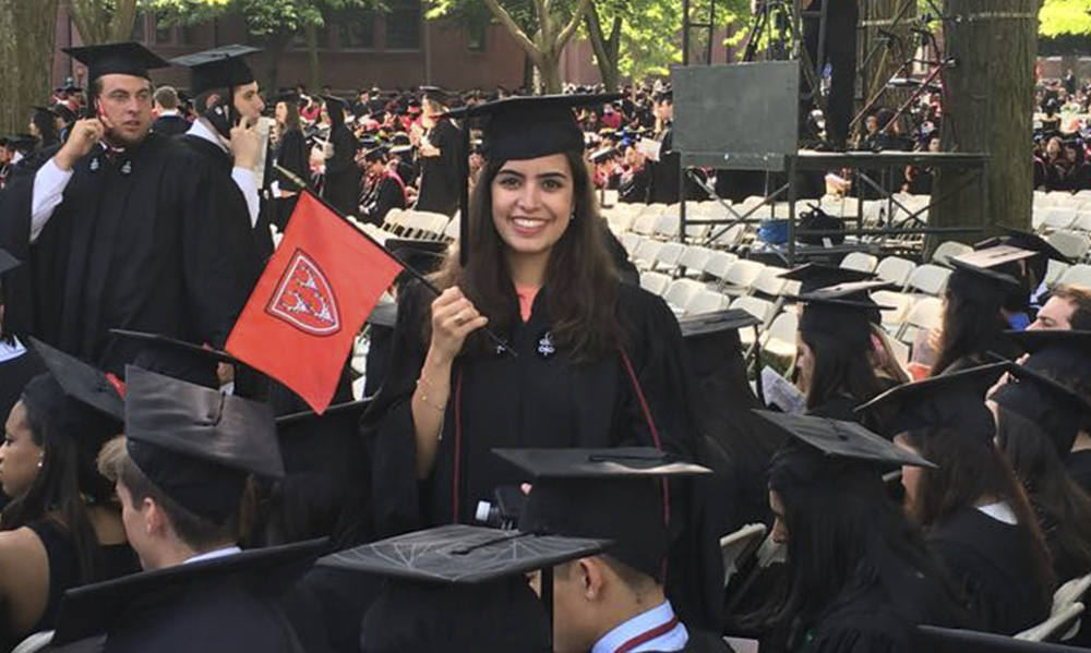 Uma bela jovem com uniforme de formando e segurando uma bandeirinha vermelha com o que parece ser o brasão da universidade de Harvard