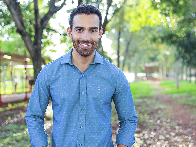 Um homem moreno e sorridente está posando para quem bateu a foto em meio a um espaço arborizado