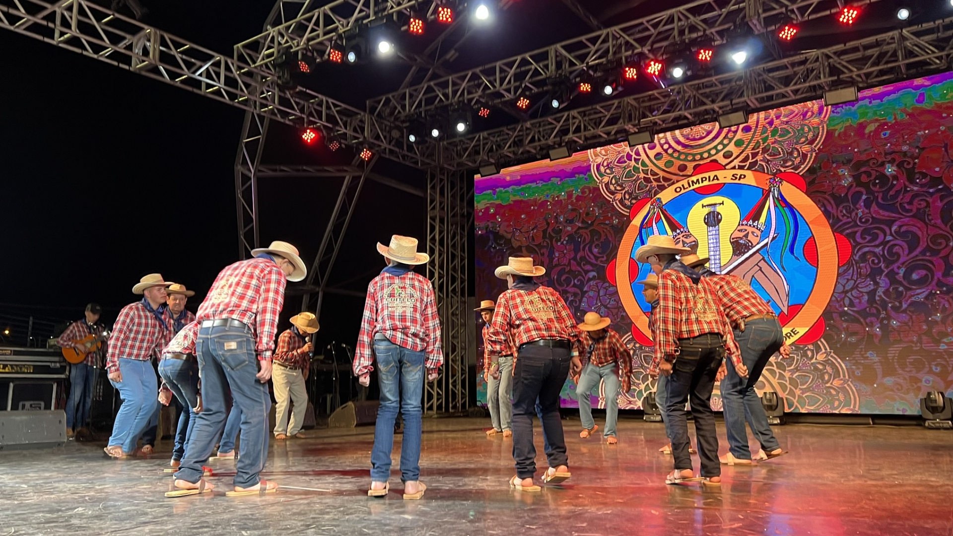 Pessoas dançam no palco. Todas usam camisas xadrez e chapéus de palha.