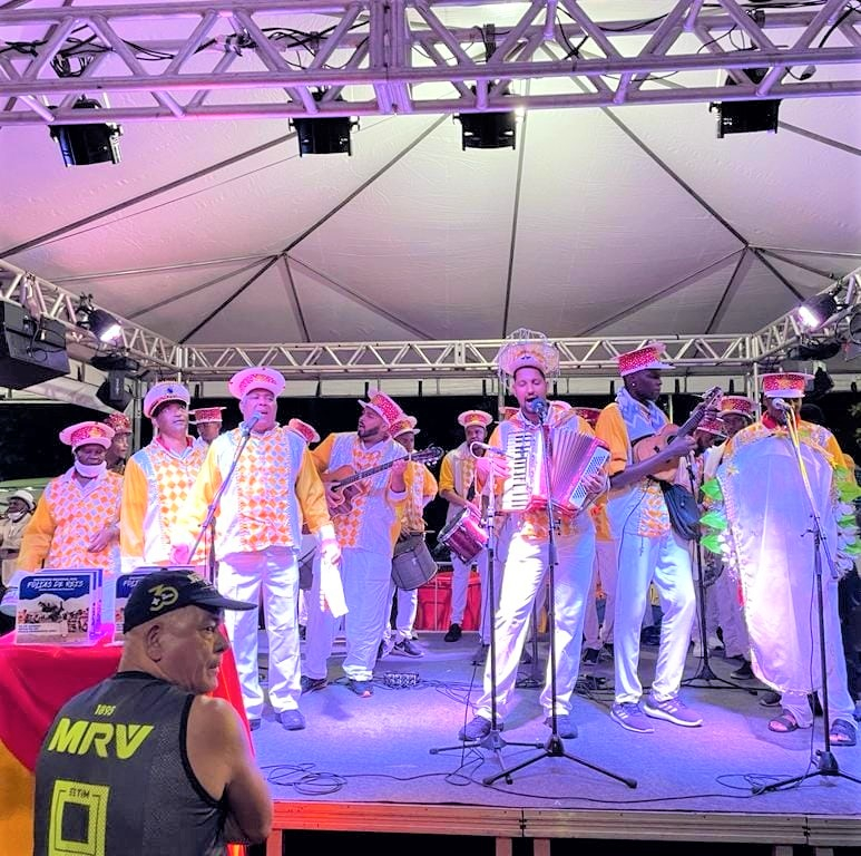 Homens vestidos de branco, com detalhes em amarelo e chapéus com detalhes em vermelho, compõem uma banda que toca em um palco também branco.