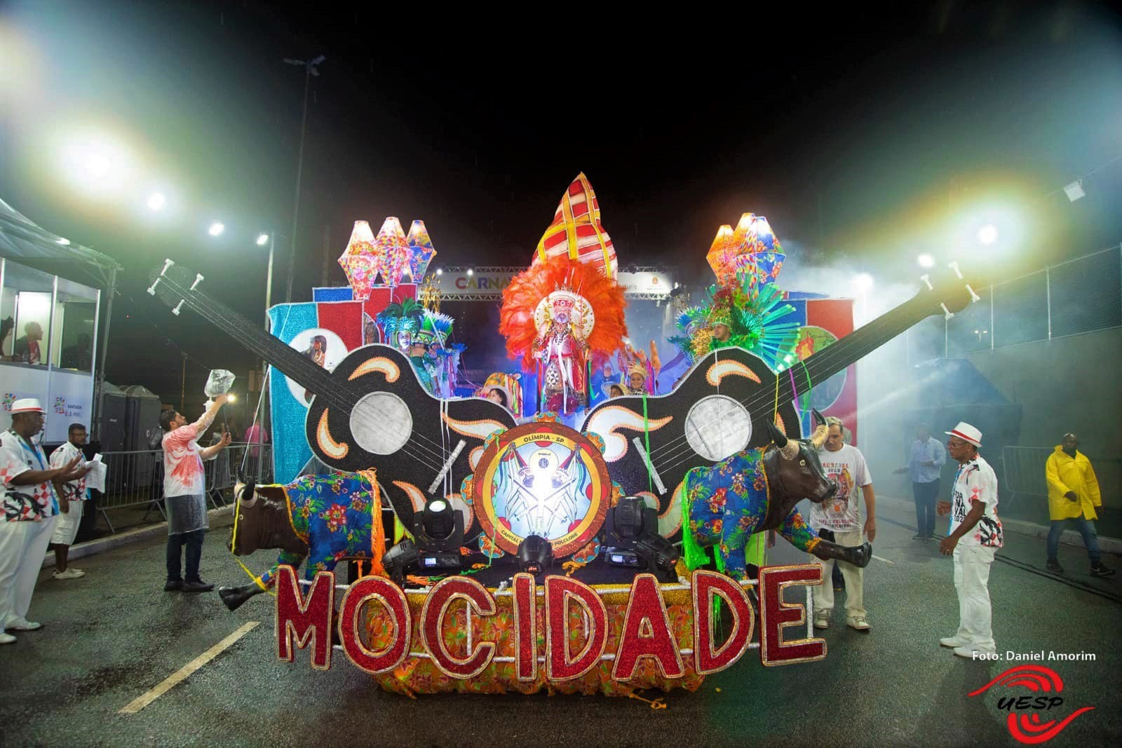 Um carro alegórico de escola de samba com a palavra "Mocidade" escrita nele. Violões e adereços coloridos também fazem parte do veículo.
