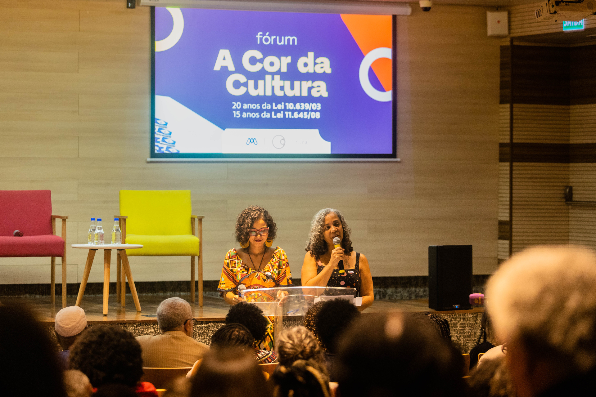 Duas mulheres estão de pé, diante de um púlpito e da plateia.