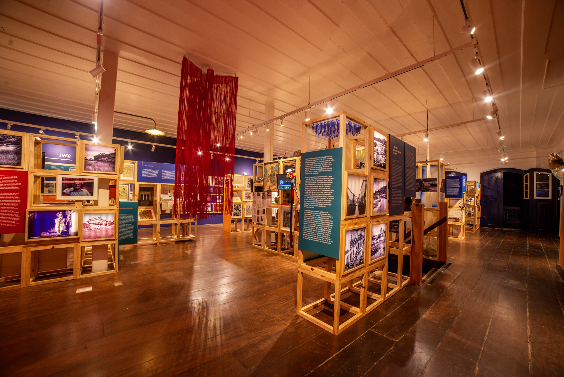 Um espaço de exposição, com chão de madeira e estantes quadradas de madeira pintada de amarelo.