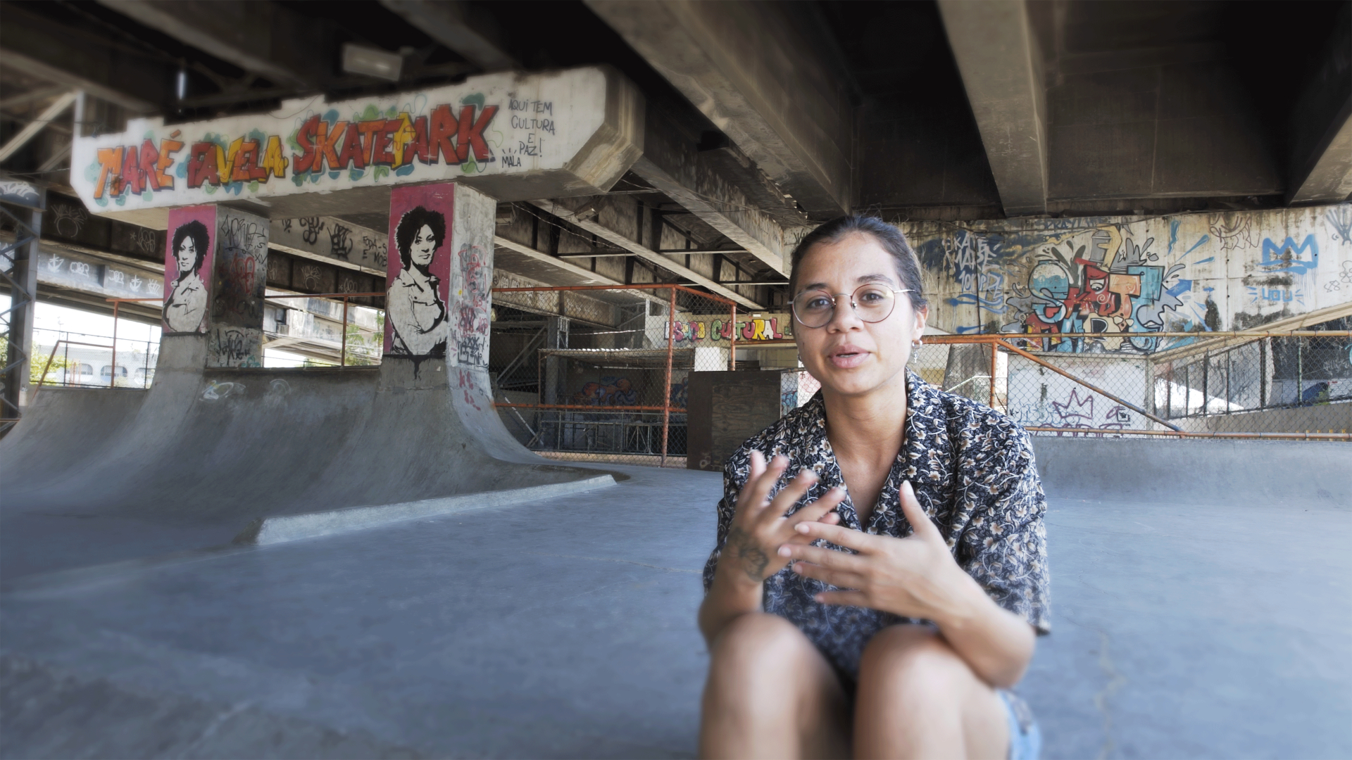 A imagem mostra uma jovem sentada próxima a um viaduto.