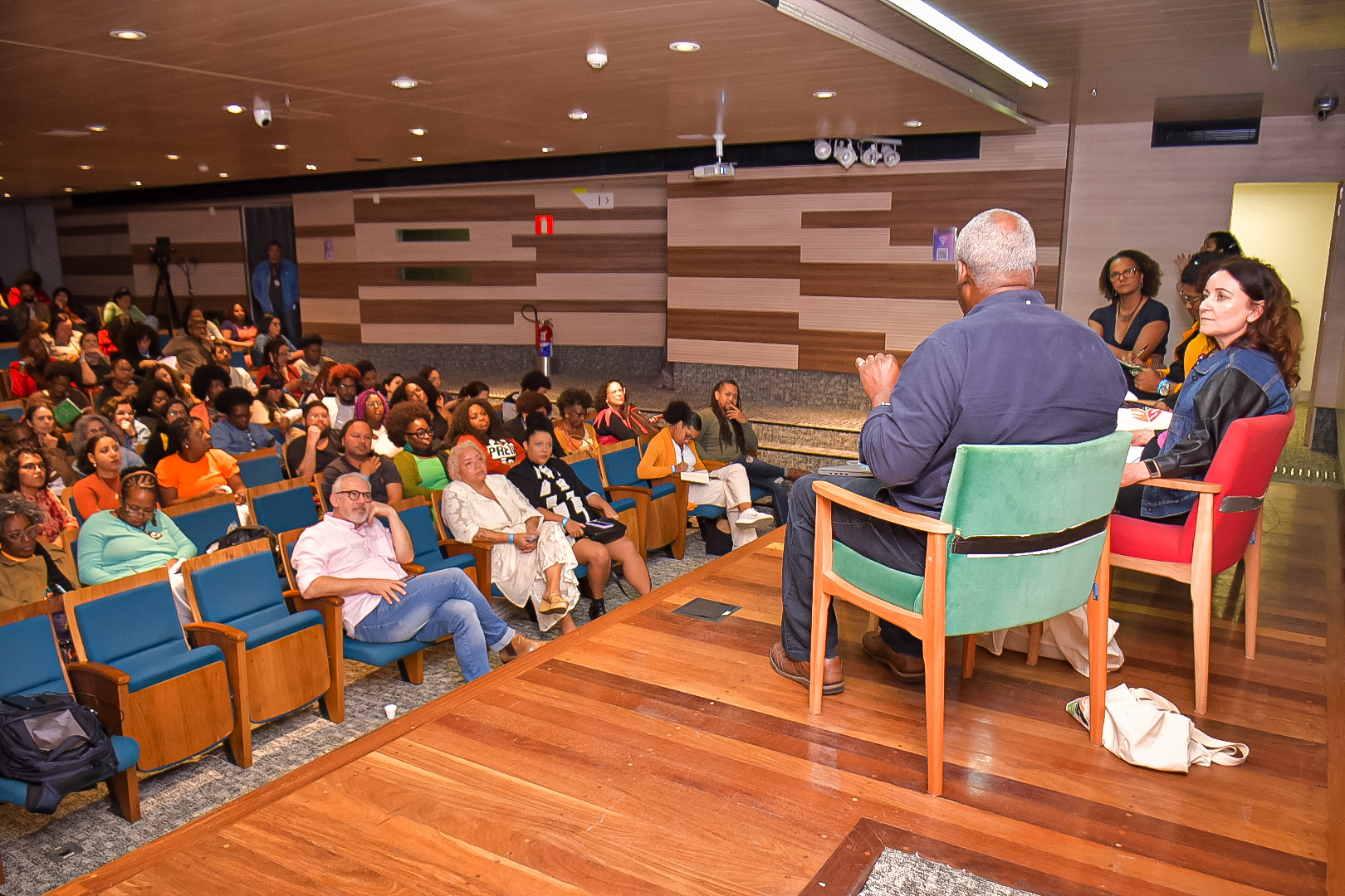 Vista de um palco de madeira em um auditório. Abaixo, o público.