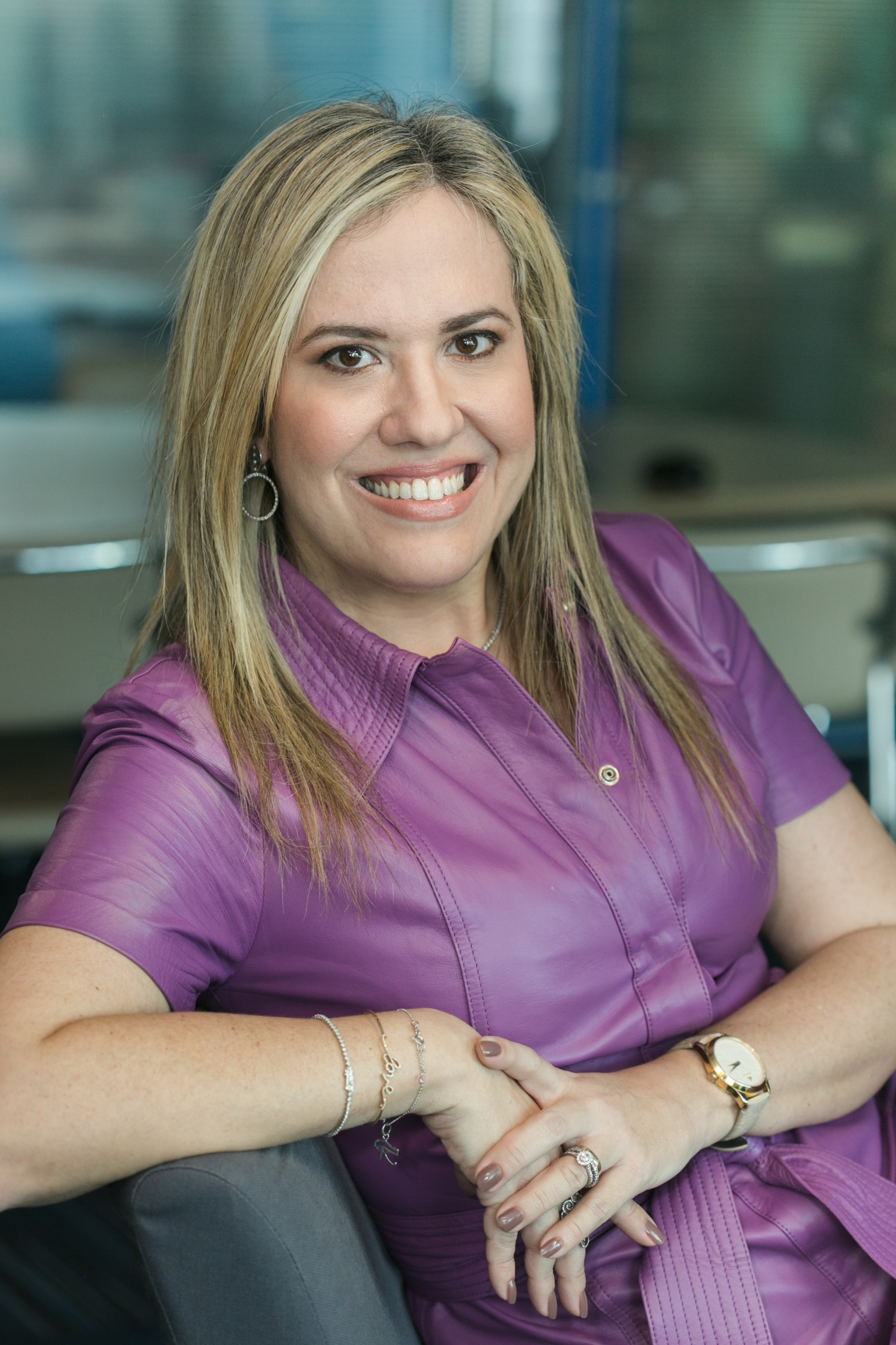 Mulher sorri. Ela está apoiada sobre o encosto de uma poltrona.