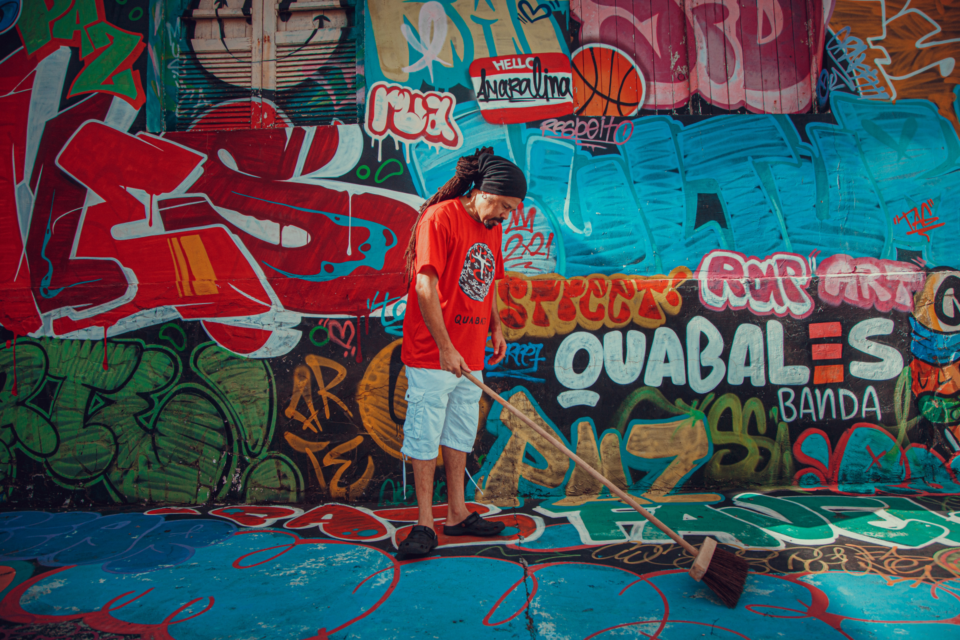 Homem negro, de cabelo comprido, varre o chão. Ao fundo, um muro pintado com grafite.
