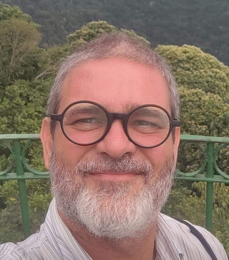 Homem sorri. Ele usa óculos redondos. Ao fundo, uma floresta.