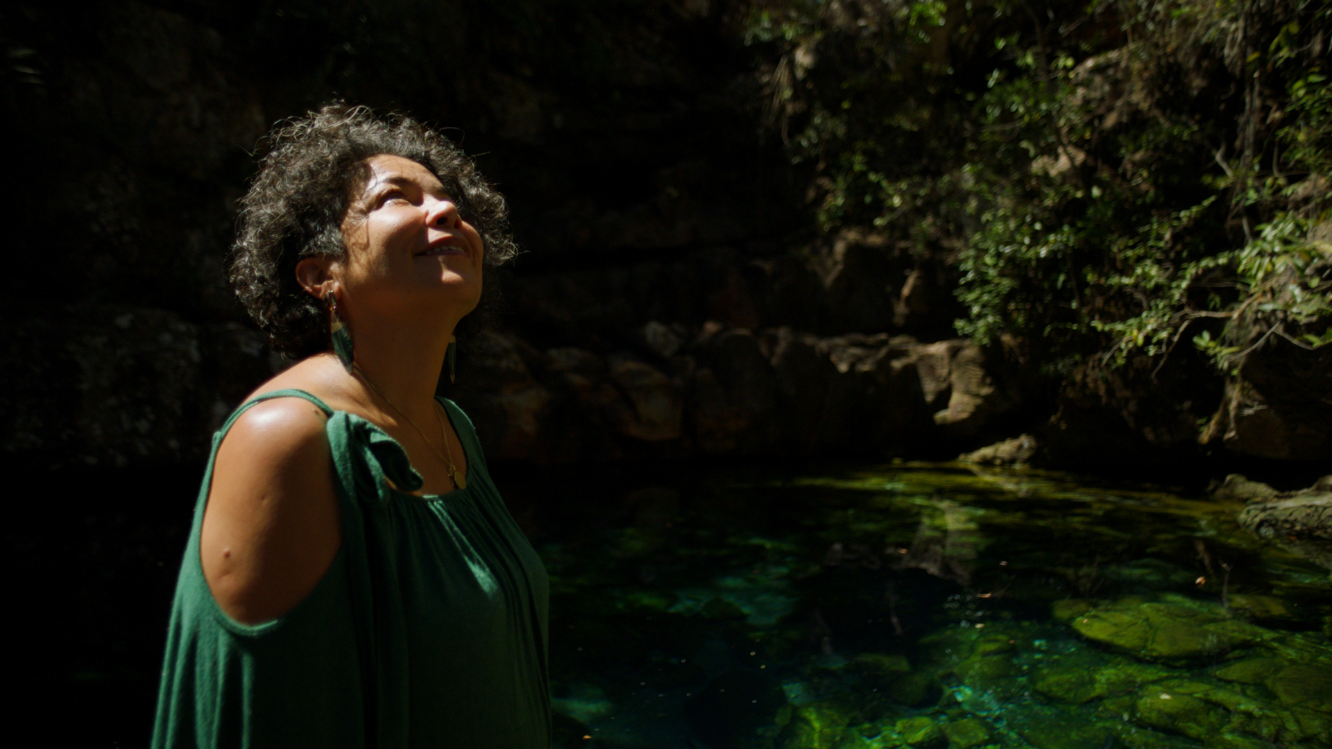 Uma mulher olha pra cima. Ao fundo, um lago, plantas e pedras.