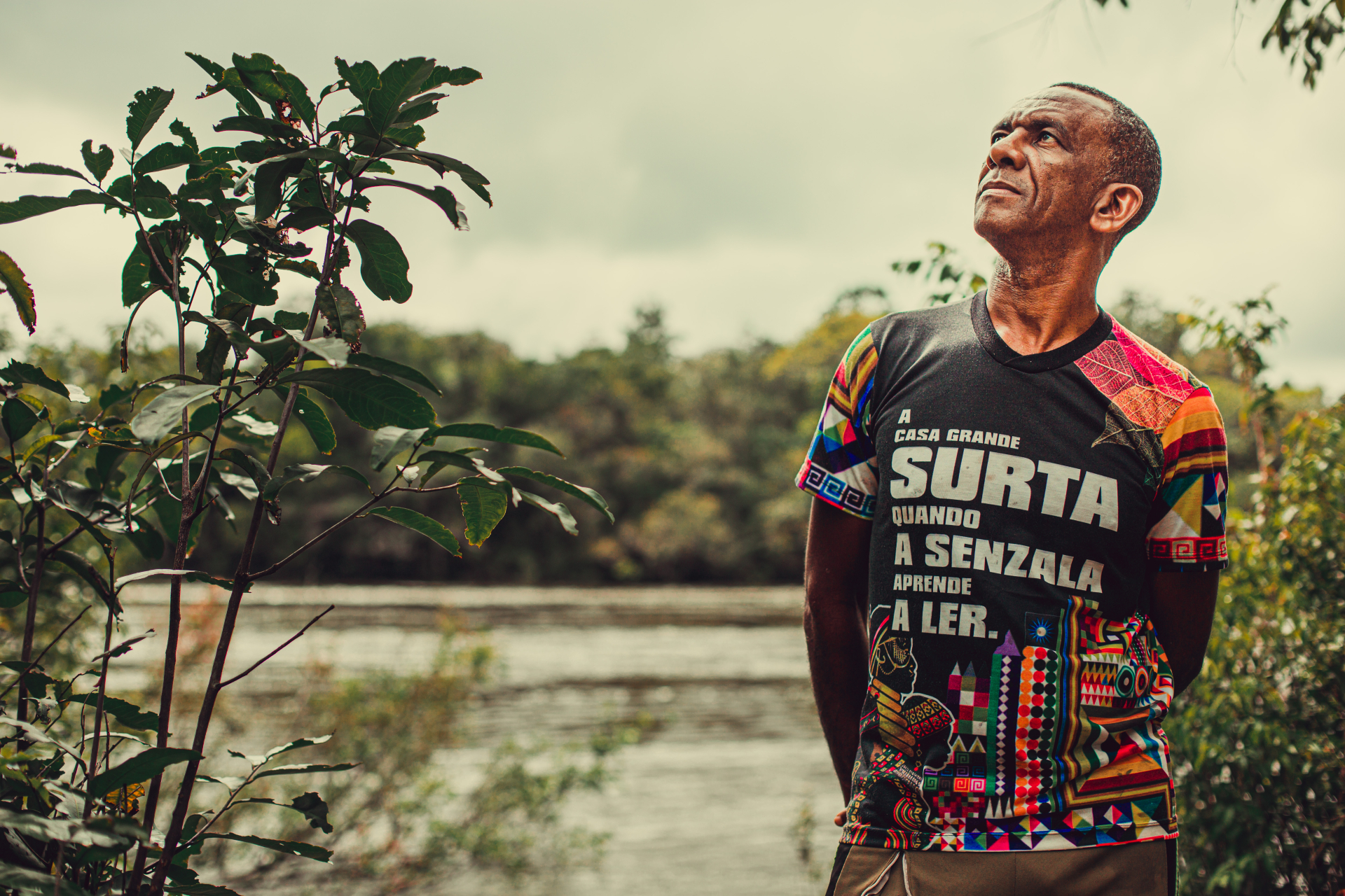 Homem negro olha para o céu. Ao fundo, um rio e a vegetação da margem.