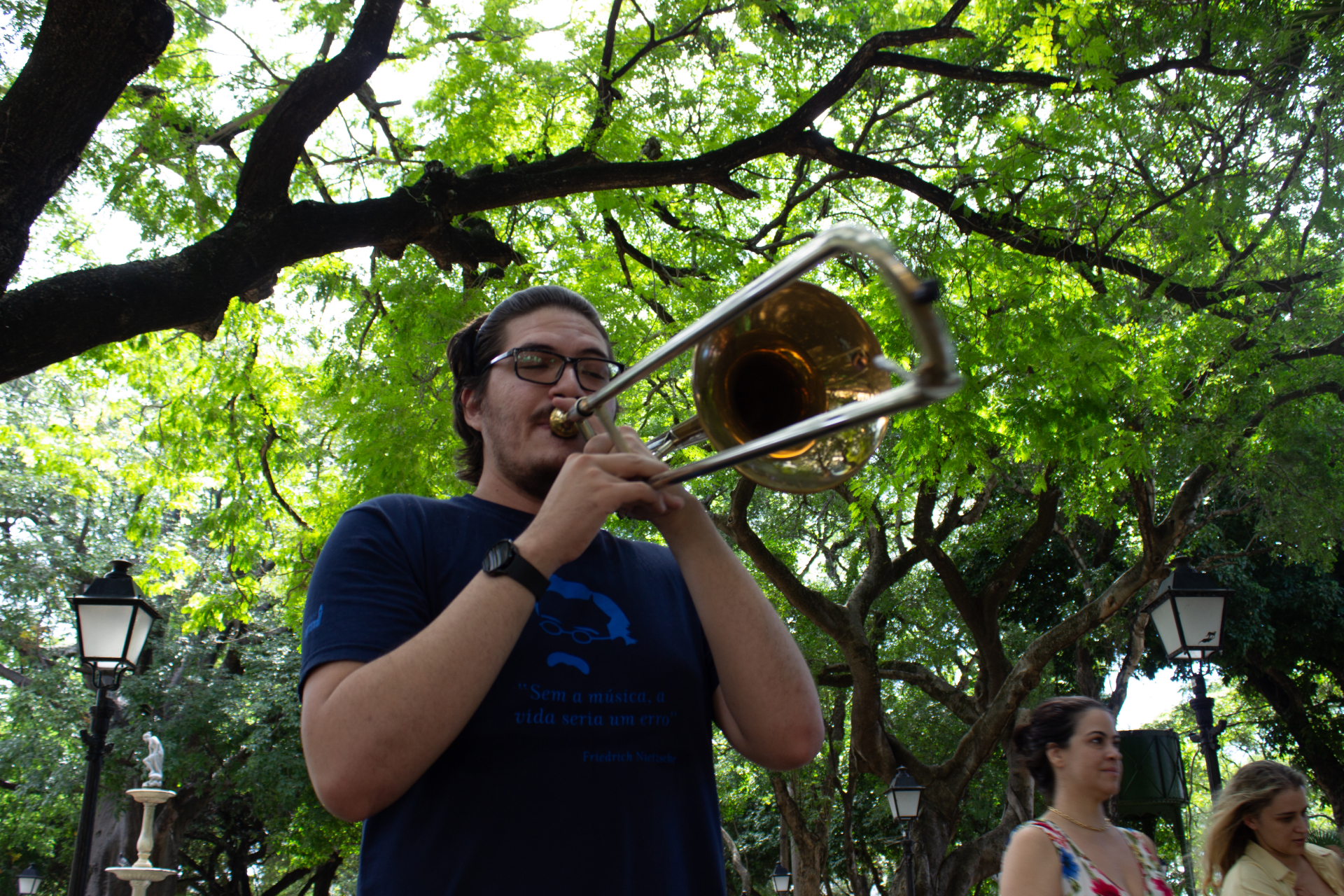 Um homem toca trombone ao ar livre. Ao fundo, árvores com folhas bem verdes.