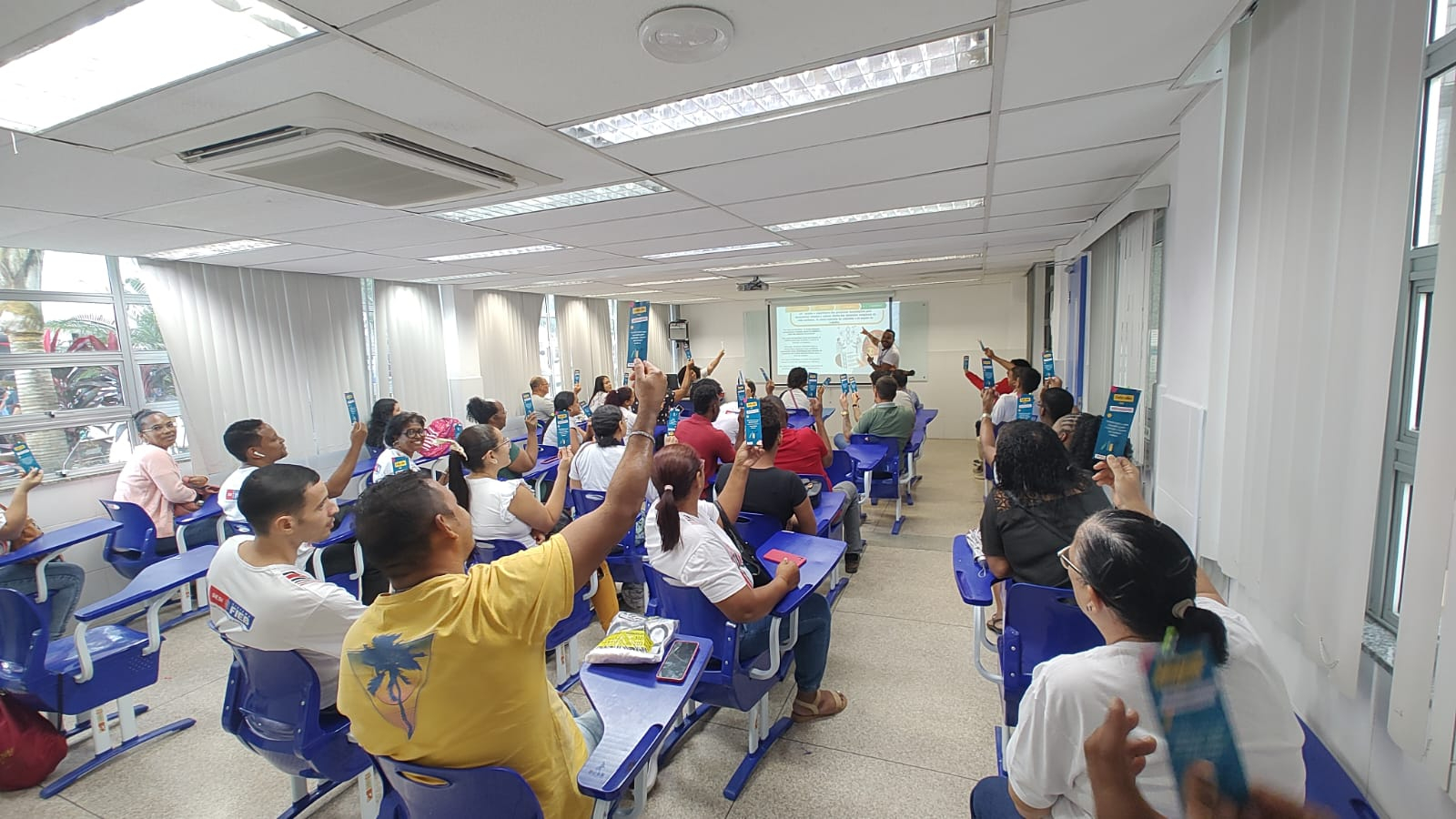 Diversas pessoas estão sentadas em suas carteiras, dentro de uma sala de aula.