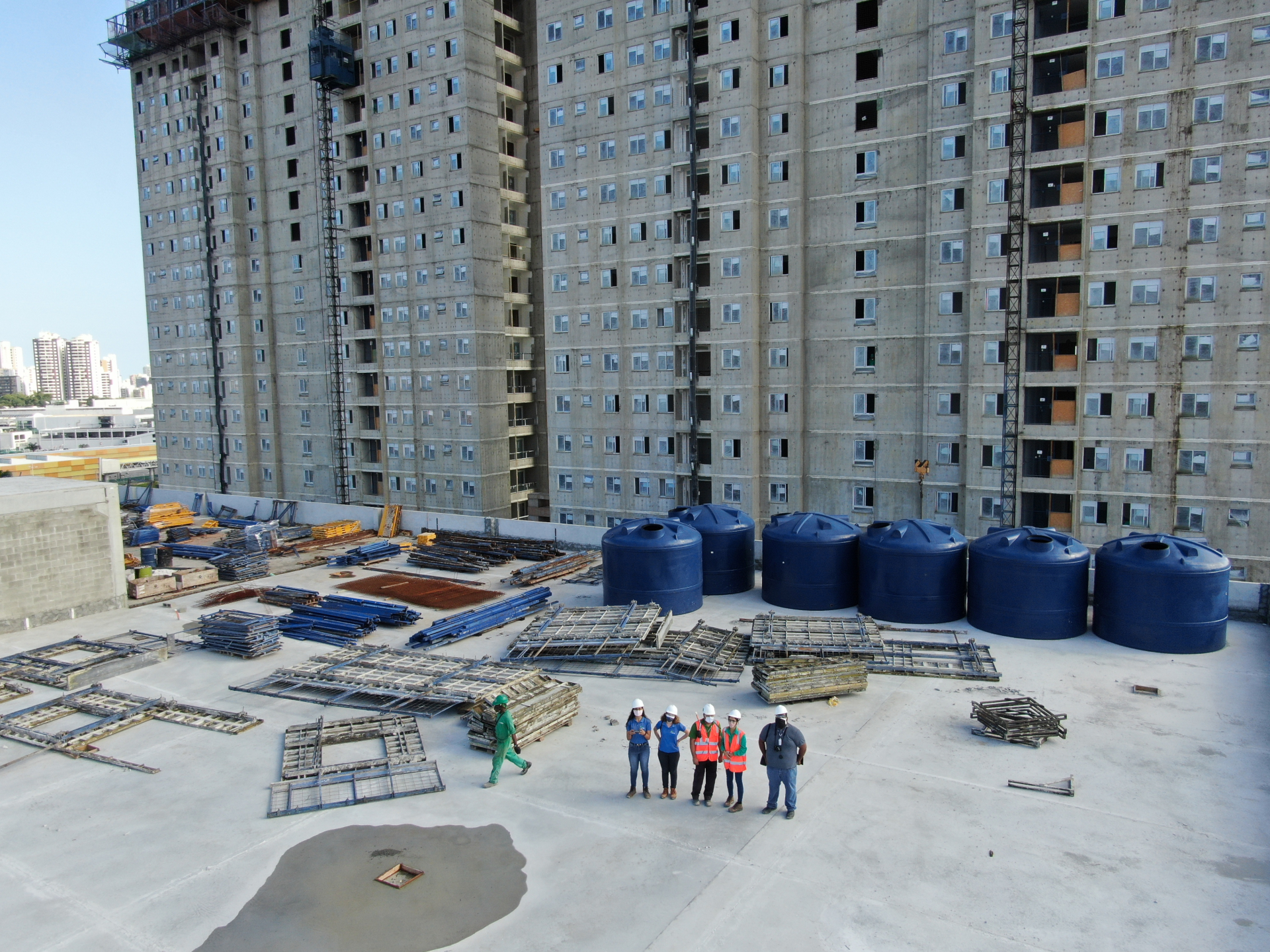 Prédios ao fundo. Diante deles, pequeninos, seres humanos estão no que parece ser um campo de obras.