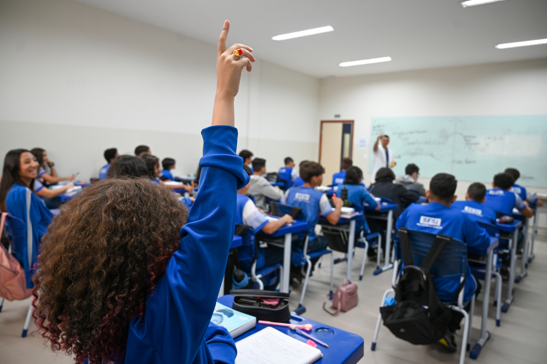 Estudantes estão sentados em suas carteiras, dentro da sala de aula. A última da fila (em primeiro plano na foto) está de mão levantada.
