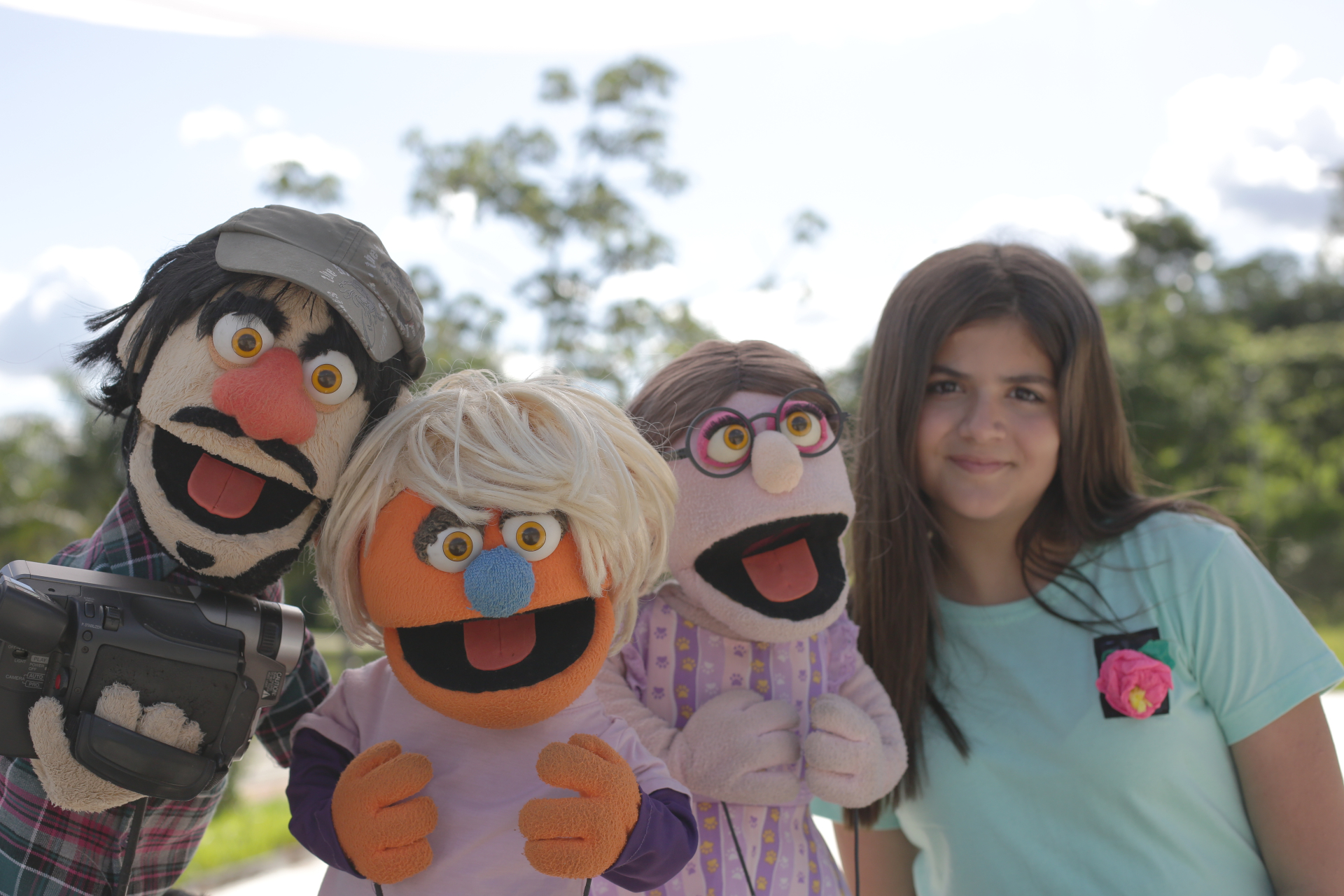 Três marionetes e uma menina sorriem. Ao fundo, árvores e o céu.