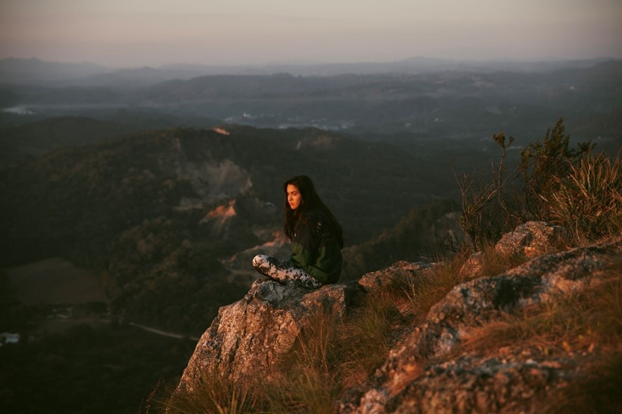 Uma mulher está sentada sobre uma pedra grande diante de uma floresta bem distante, abaixo da montanha em que ela está.