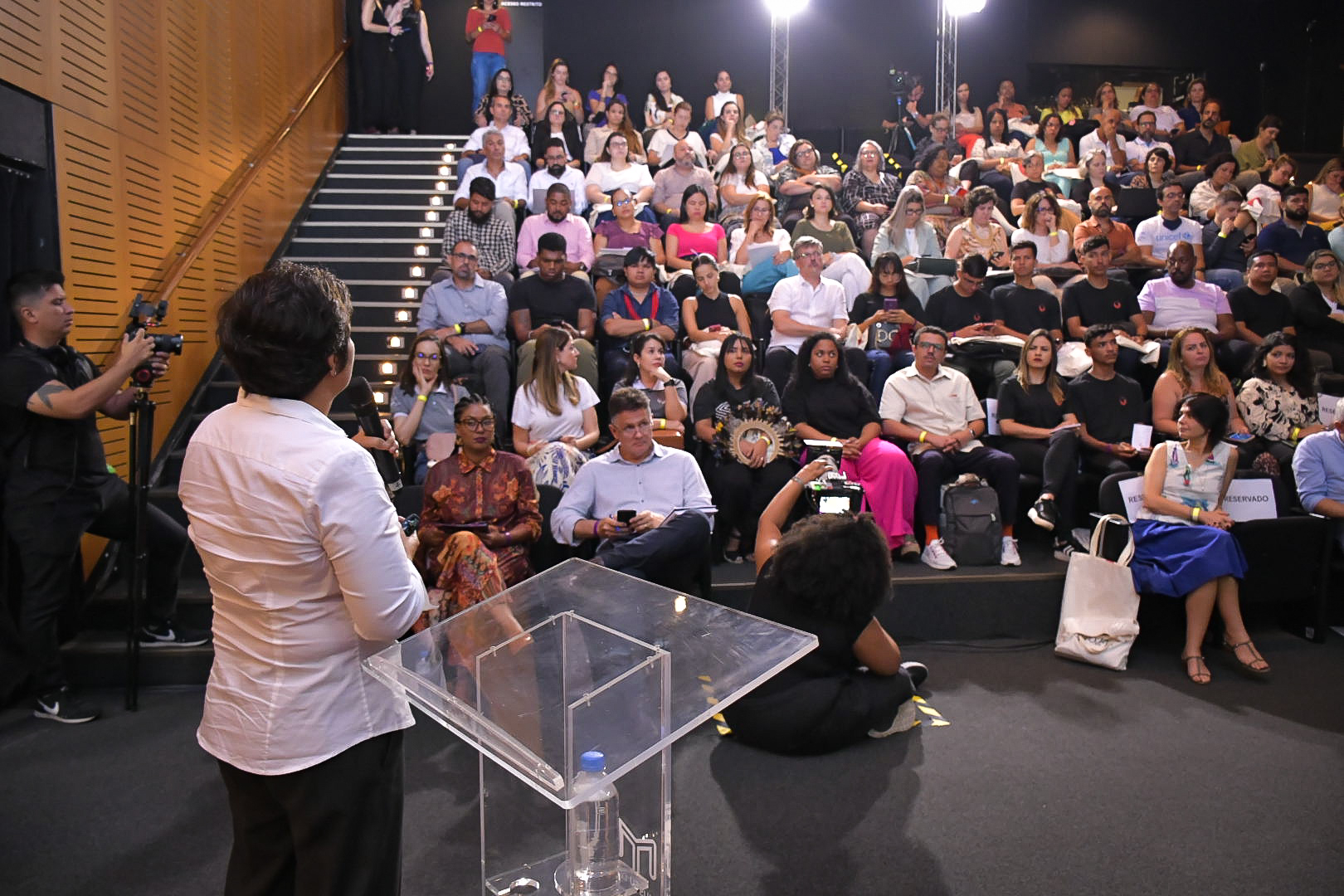 Foto tirada do fundo do palco para o auditório. No palco, uma mulher fala ao microfone. Ao fundo, o público a assiste.