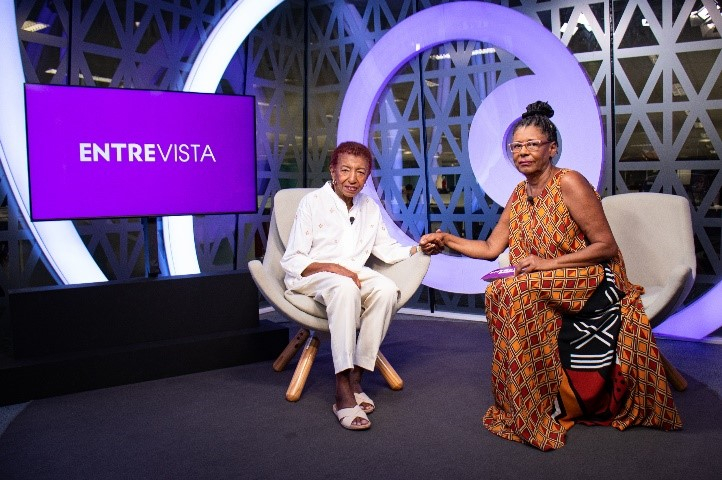 Duas mulheres negras, de mãos dadas, estão sentadas em poltronas claras. Ao fundo, uma tv e decoração em tons de lilás.