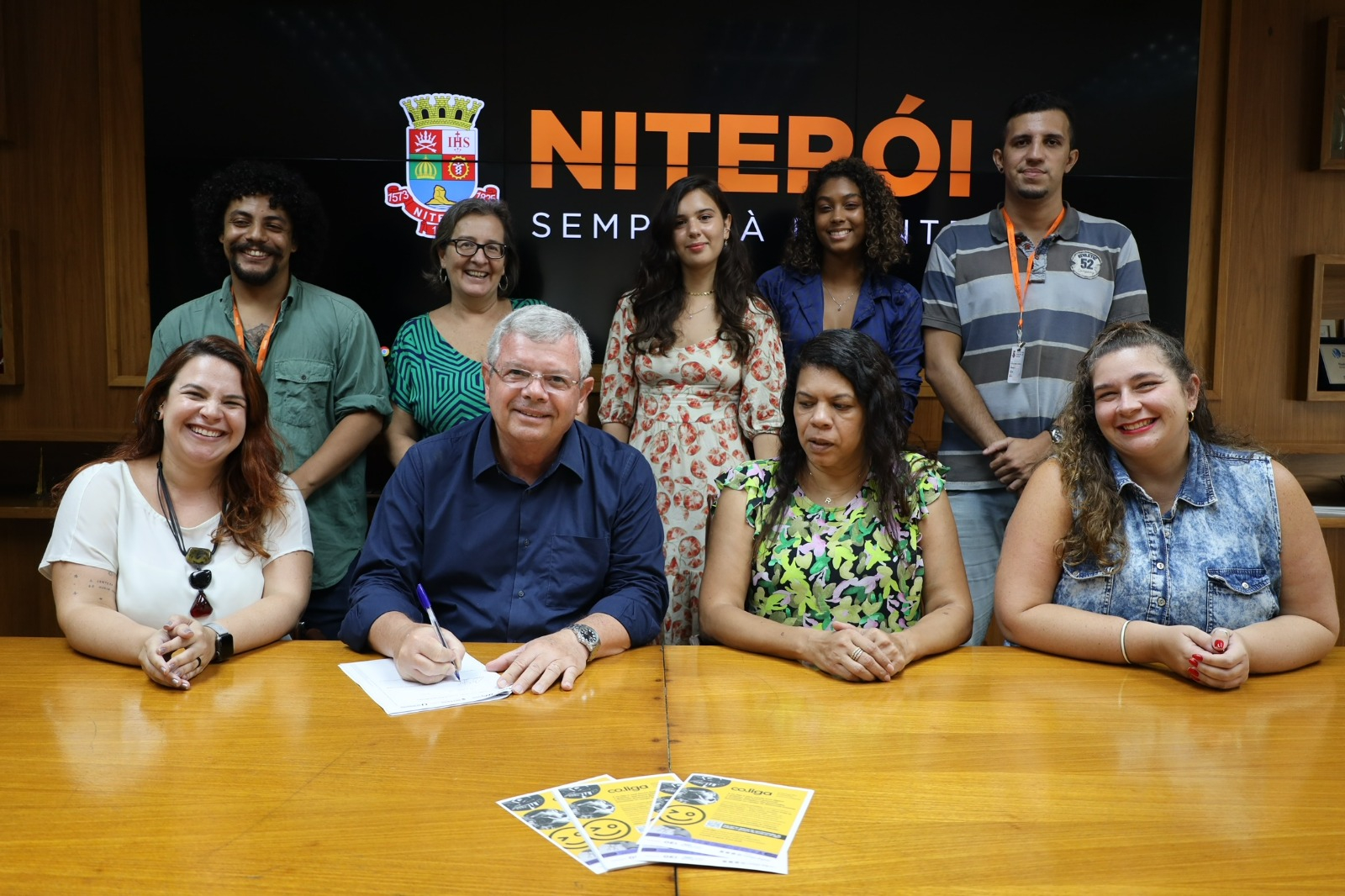 Cinco pessoas estão de pé, atrás de quatro pessoas sentadas. Todas estão diante de uma grande mesa de madeira em tom amarelo. Ao fundo, uma faixa com a palavra "Niterói".