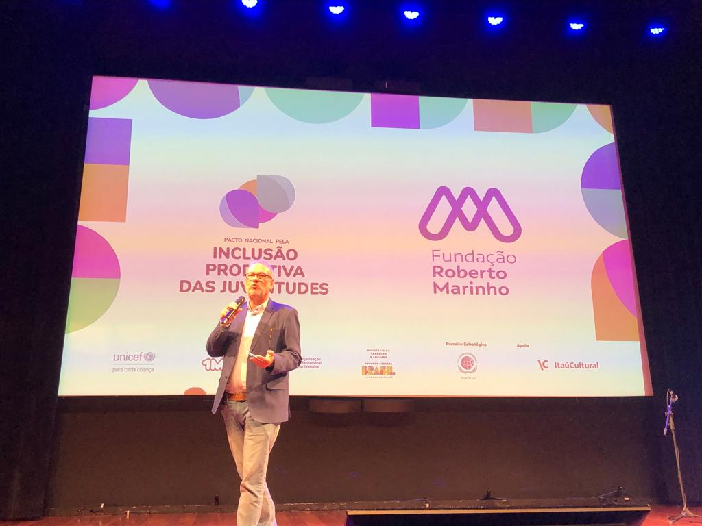 Um homem caminha sobre um palco, com um microfone na mão. Ao fundo, um telão de fundo branco tem a logomarca da Fundação Roberto Marinho e do Pacto Nacional pela Inclusão Produtiva das Juventudes.
