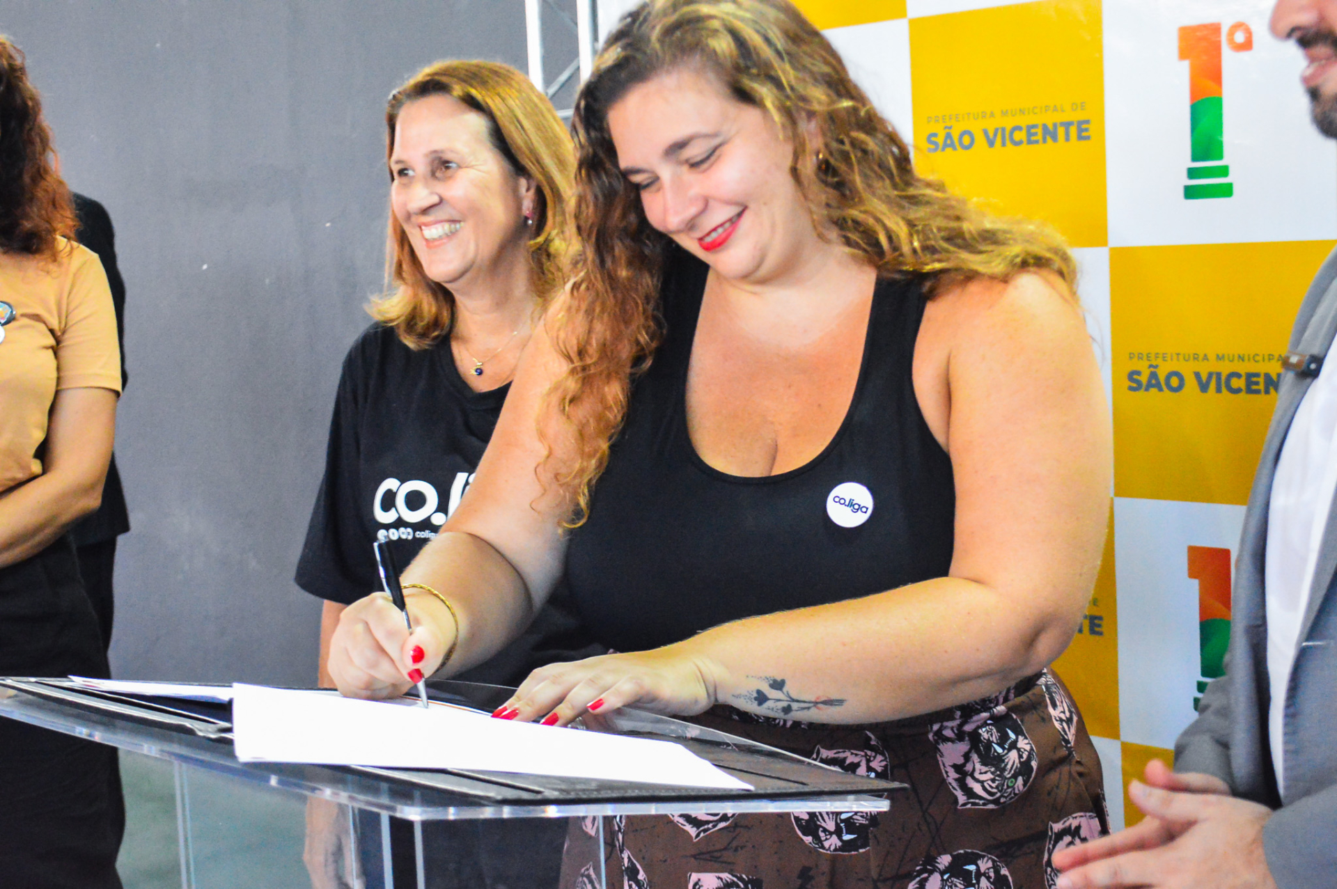 Mulher está de pé, sorrindo, enquanto assina um papel que está sobre um púlpito.