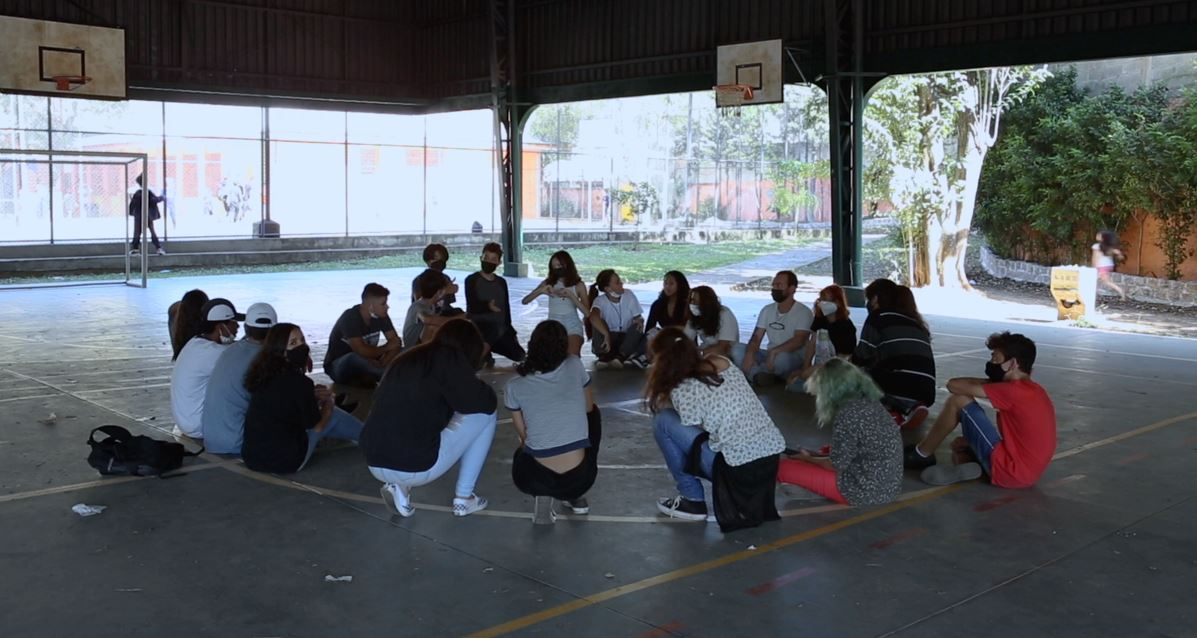 Jovens estão sentados e sentadas no chão de uma quadra de esportes.