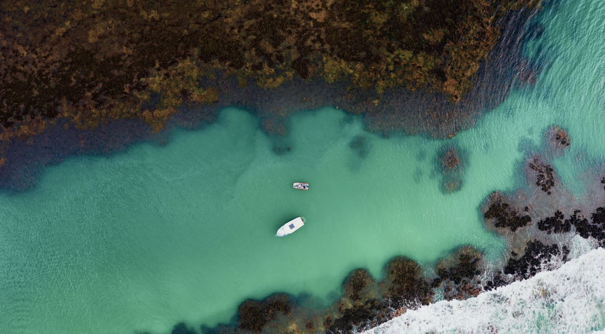 Fotografia feita de cima, mostrando o mar em azul bem claro, com um pequeno barco navegando por ele. Ao redor, vegetação em verde bem escuro nas margens.
