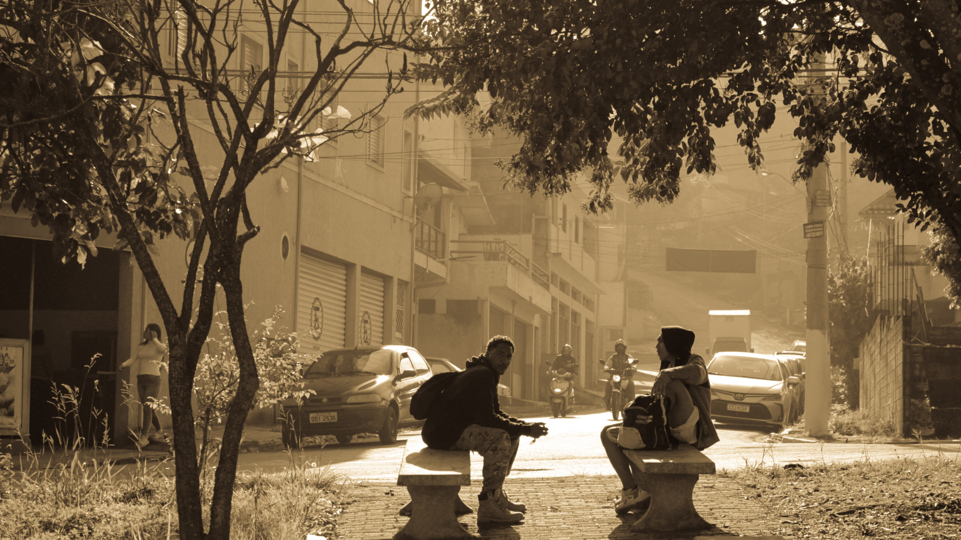 Duas pessoas estão sentadas em bancos de um local público, a céu aberto. Há árvores ao redor deles e a cena está em tons de sépia (como um preto e branco, mas envelhecido)