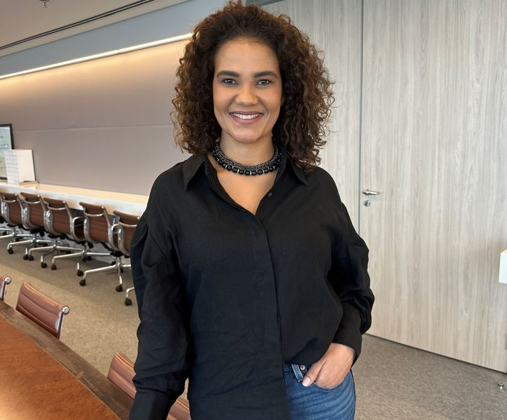Uma mulher negra sorri, de pé, com uma mão no bolso da calça jeans que veste. Ao fundo, a parede e algumas cadeiras do auditório em que ela está.