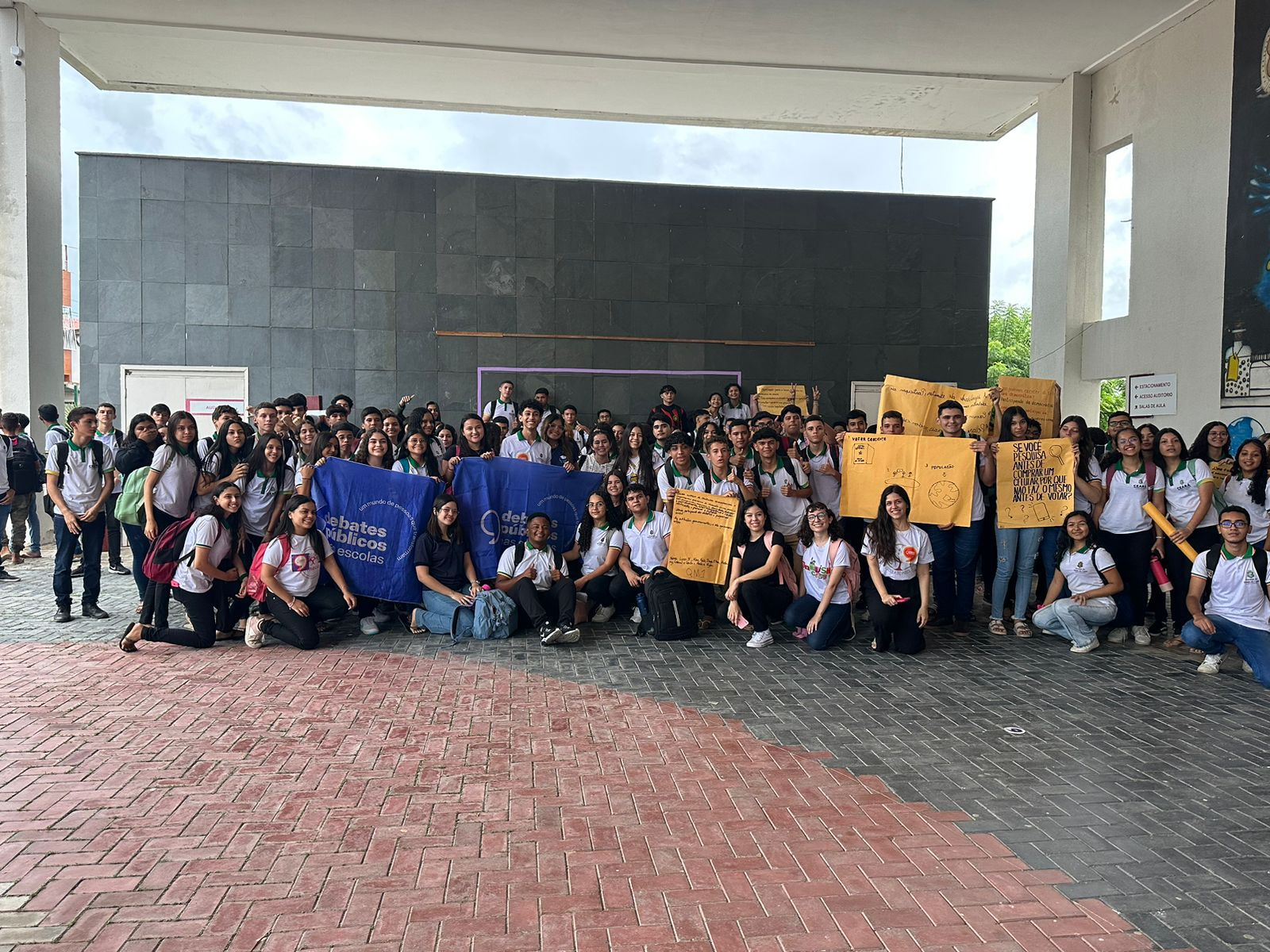 Jovens reunidos sorrindo, de pé, segurando uma bandeira em que está escrito "Debates públicos nas escolas".