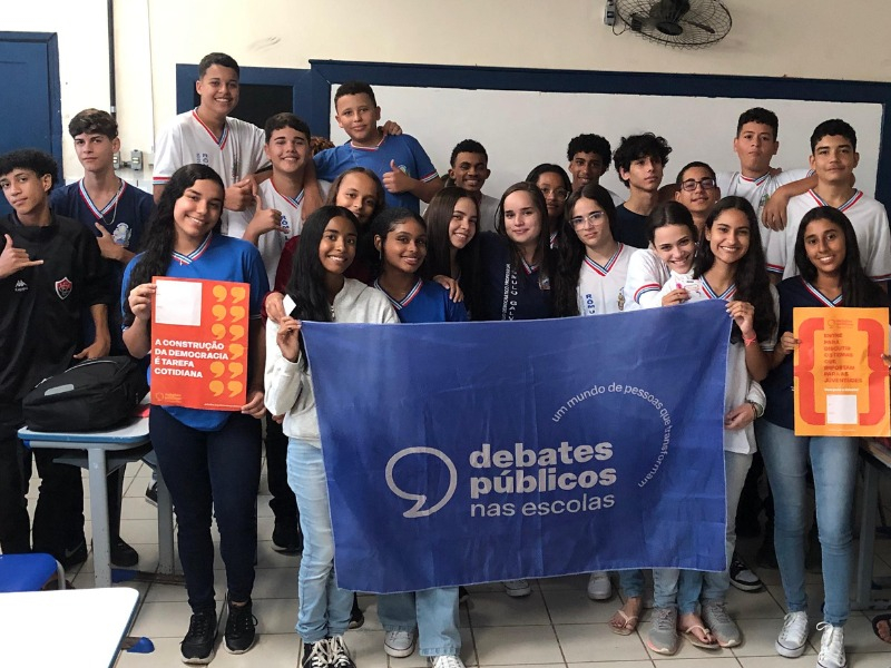 Jovens reunidos sorrindo, de pé, segurando uma bandeira em que está escrito "Debates públicos nas escolas".