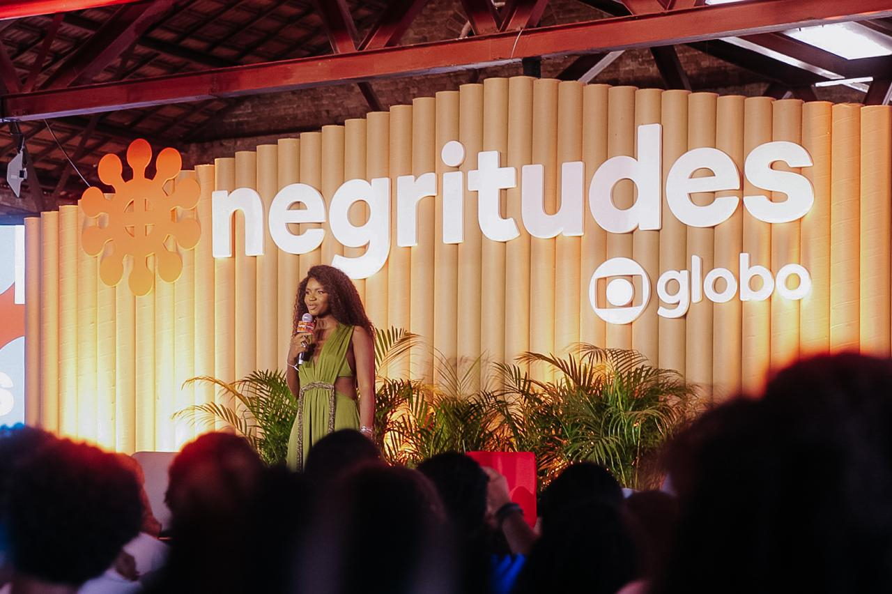 Uma mulher negra fala ao microfone diante de uma plateia.  Ao fundo, o nome do festival "Negritudes Globo".