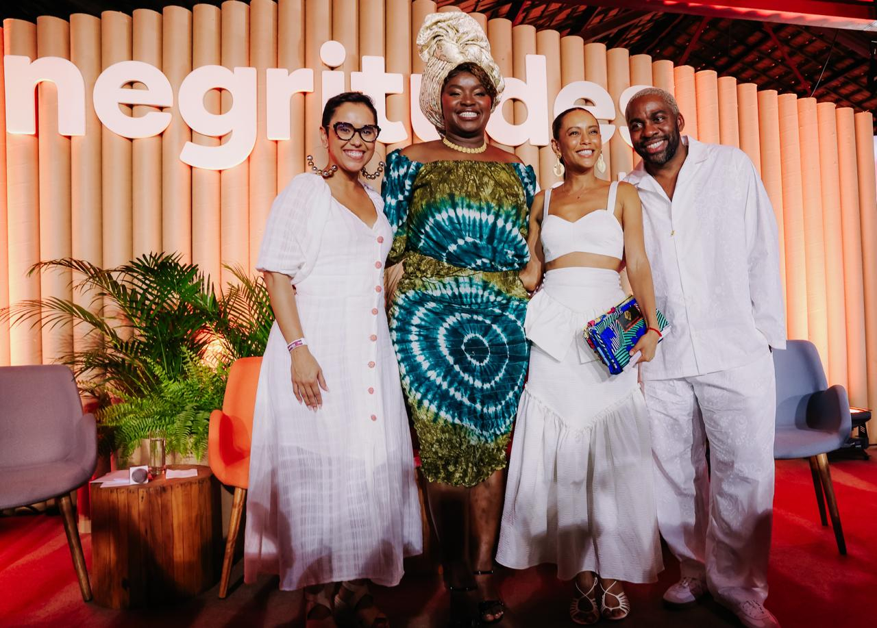 Quatro pessoas negras estão de pé. Ao fundo, o nome do evento: "Negritudes".