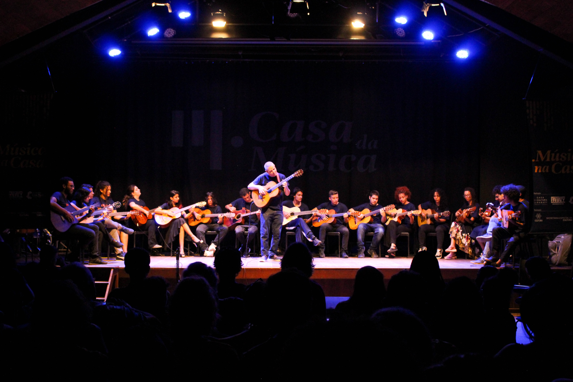 Diversas pessoas estão com violões em um palco. A fotografia foi feita a partir do público, que aparece em silhuetas.