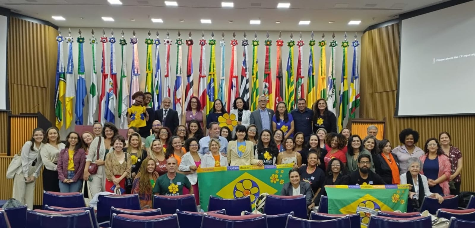 Diversas pessoas estão de pé em um palco,. sorrindo. A decoração ao fundo do palco é de bandeiras coloridas.