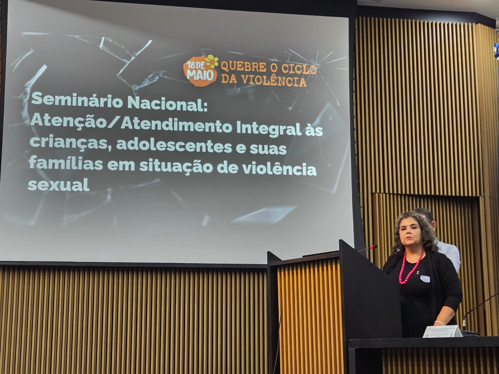 Uma mulher está de pé, diante de um púlpito. Ao seu lado, um telão com texto projeto.