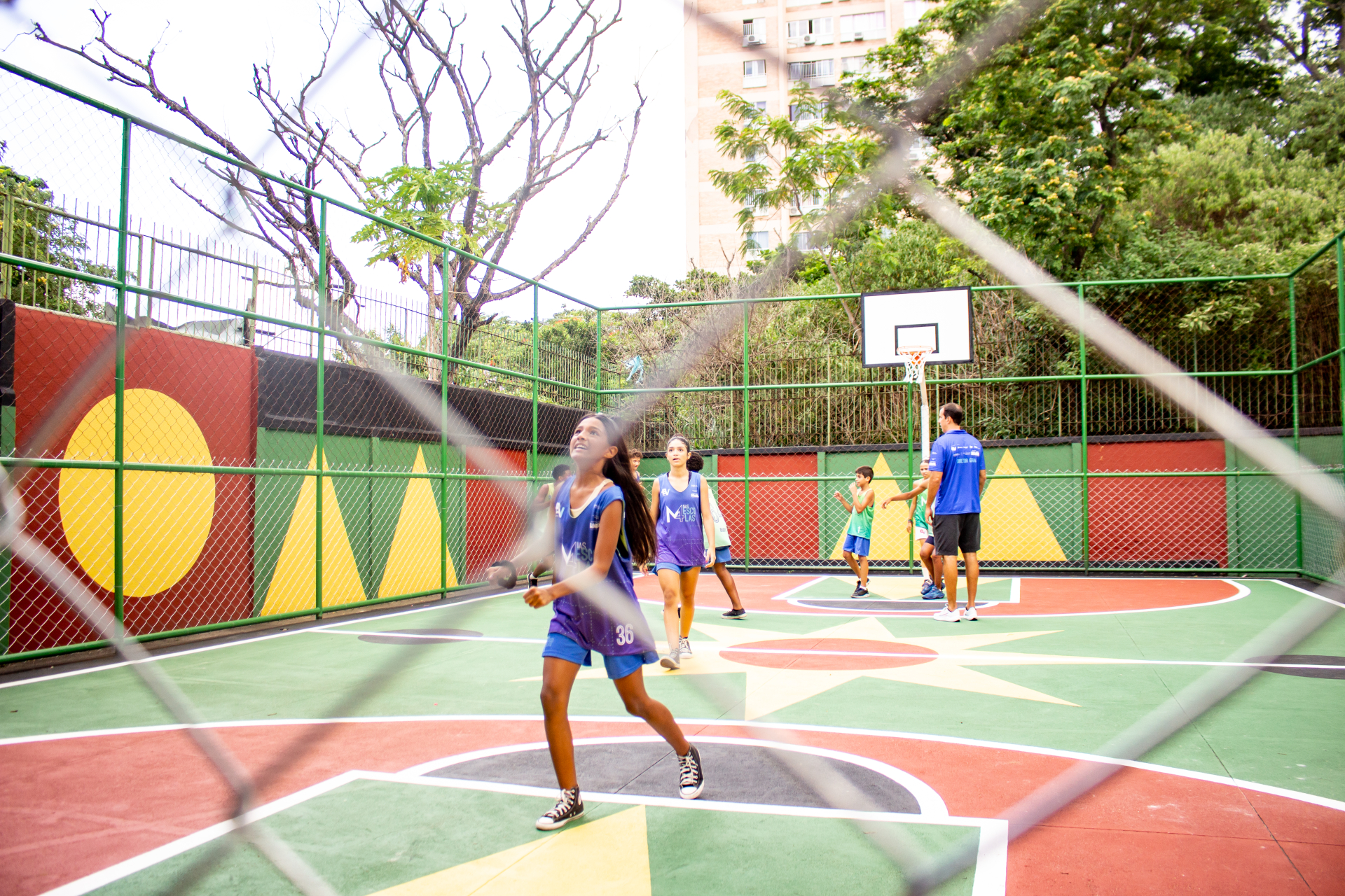 Foto mostra uma quadra de esportes colorida em que poucos jovens jogam basquete.