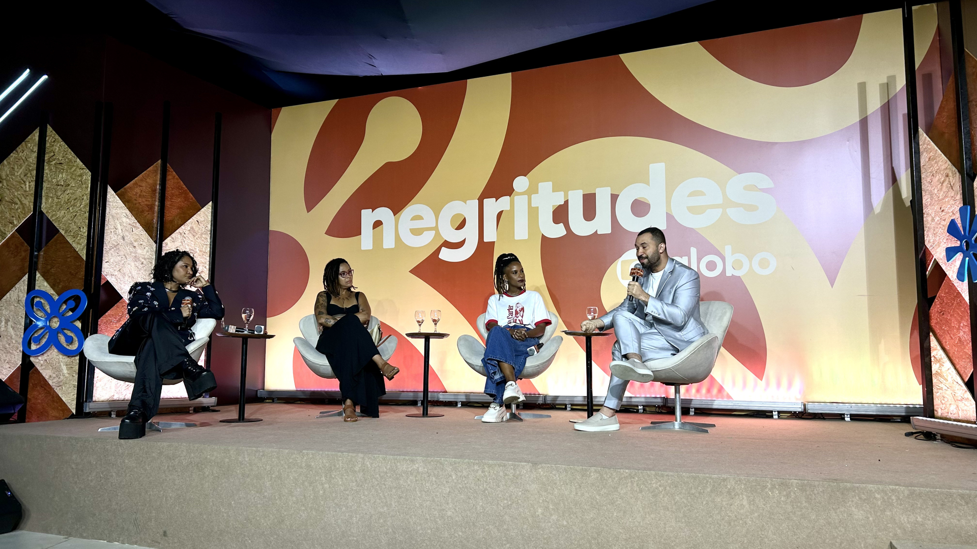 Quatro pessoas negras sentadas em poltronas sobre um palco. Ao fundo, o nome "Negritudes".
