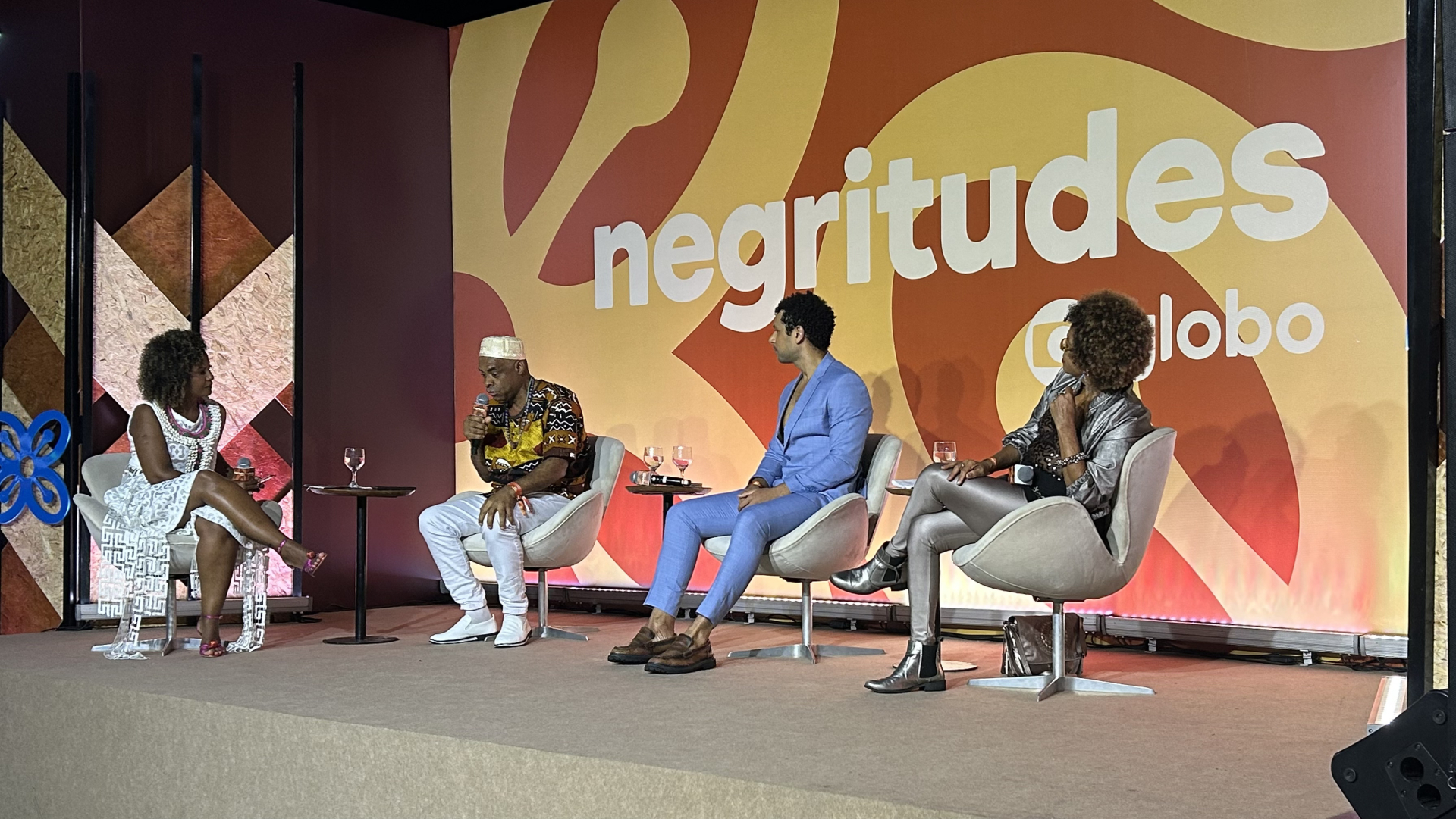 Quatro pessoas negras estão sentadas em poltronas sobre um palco, diante de uma plateia. Ao fundo, sobre uma parede em tons de marrom-claro, o nome do evento: Negritudes Globo.