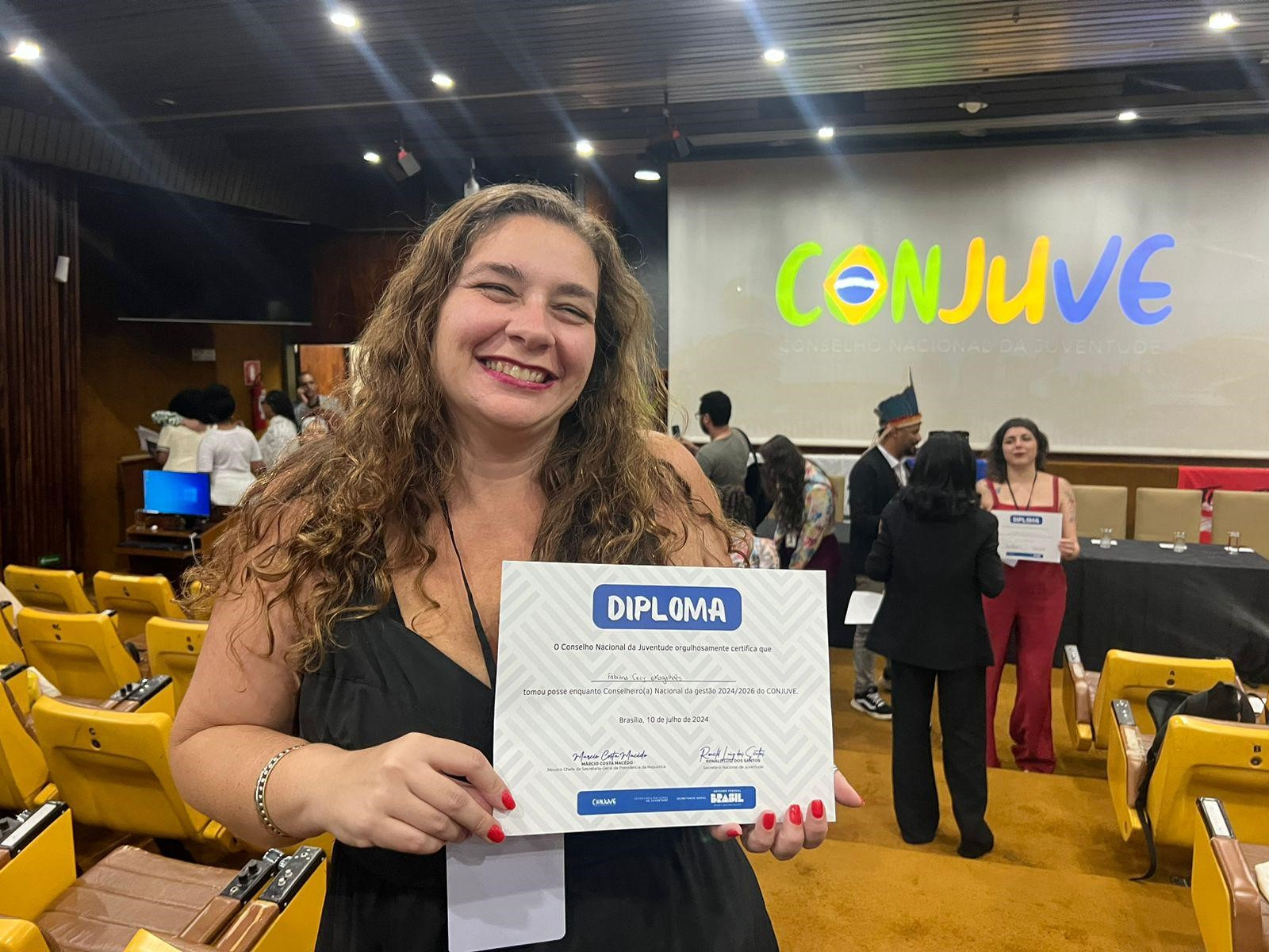 Com um auditório ao fundo (e o nome "Conjuve" projetado), uma mulher de cabelo comprido e ondulado sorri segurando um diploma.