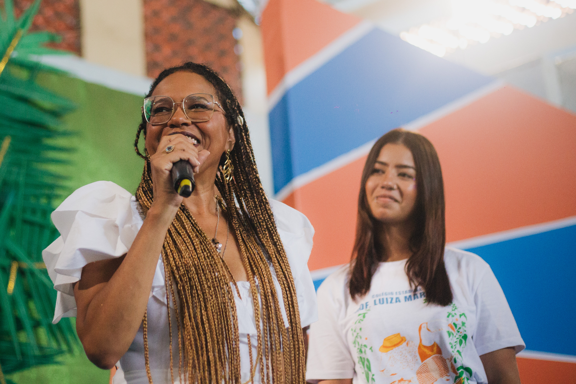 Teresa Cristina, cantora de samba, fala sorrindo ao microfone. Ao seu lado uma jovem sorri.