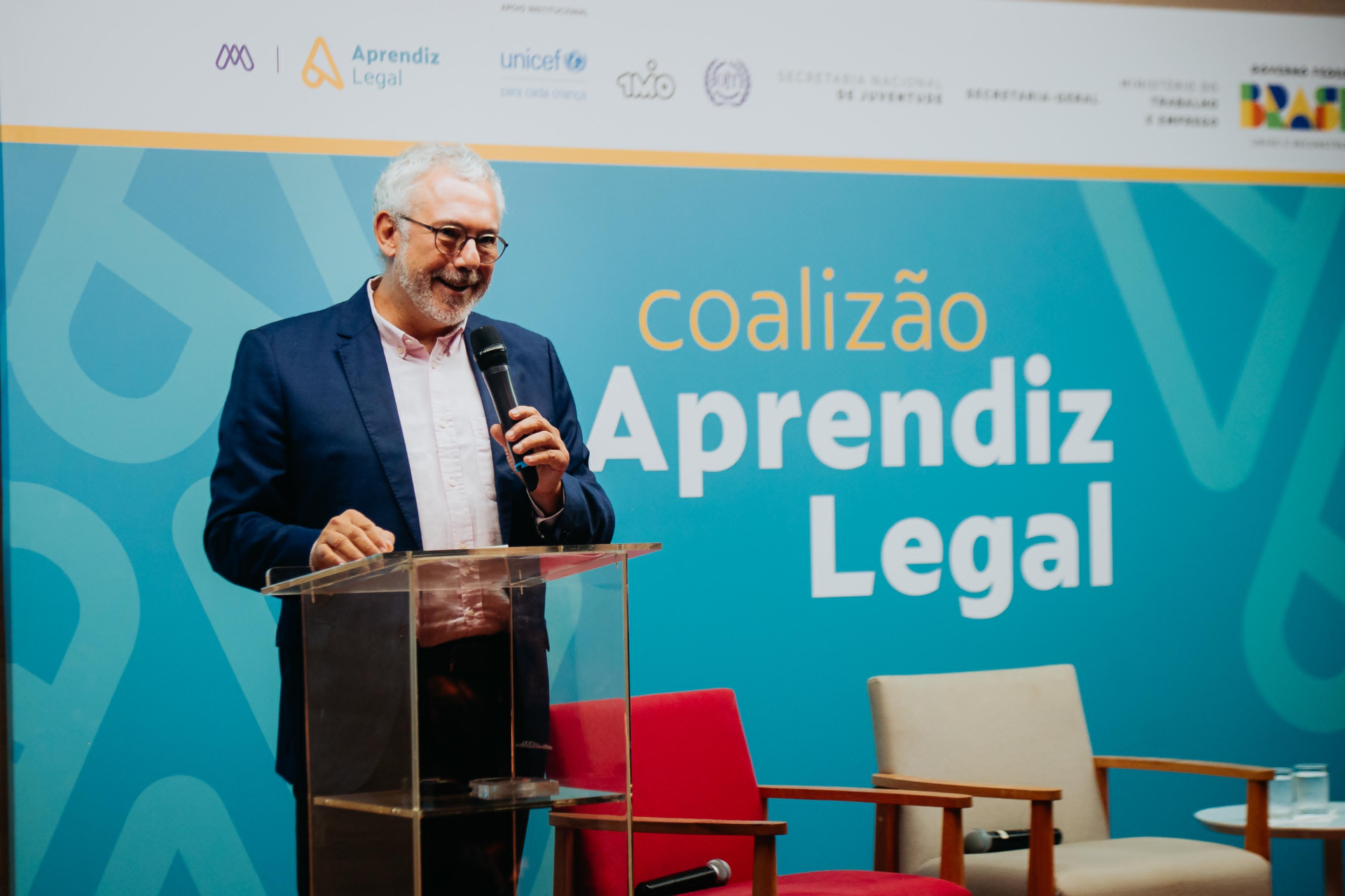 Um homem branco, com cabelo branco e óculos, sorri com o microfone na mão. Ao fundo, uma tela em azul com o texto "Coalização Aprendiz Legal".