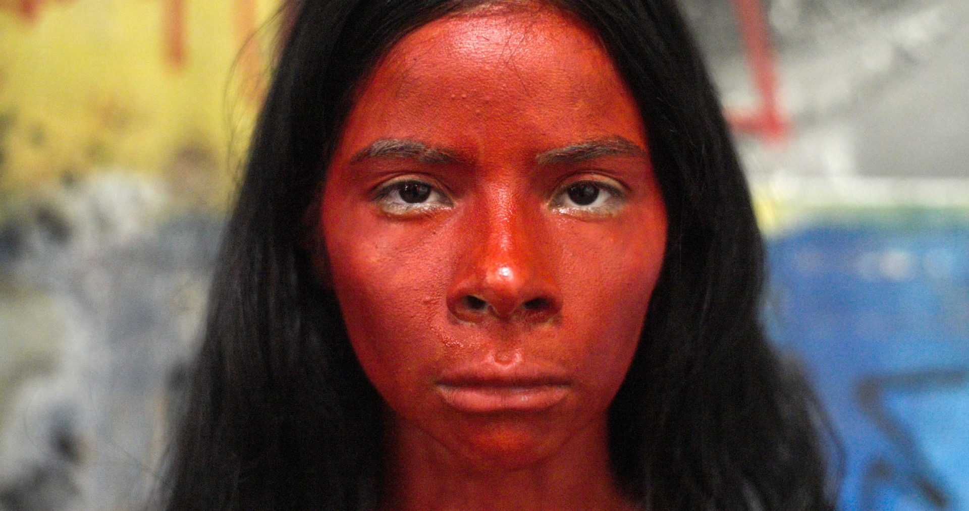 Jovem indígena, com pele pintada em tom avermelhado e cablo preto liso, olha fixamente para quem a fotografa.