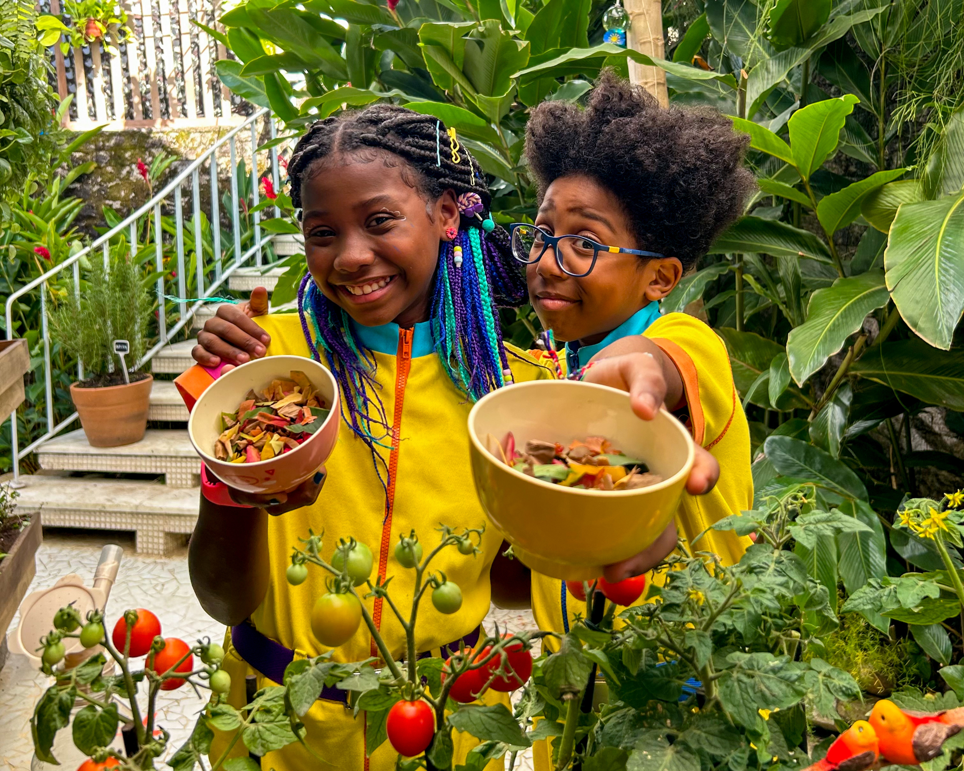 Duas crianças sorriem segurando cestos com folhas coloridas. Ambas estão em uma horta.