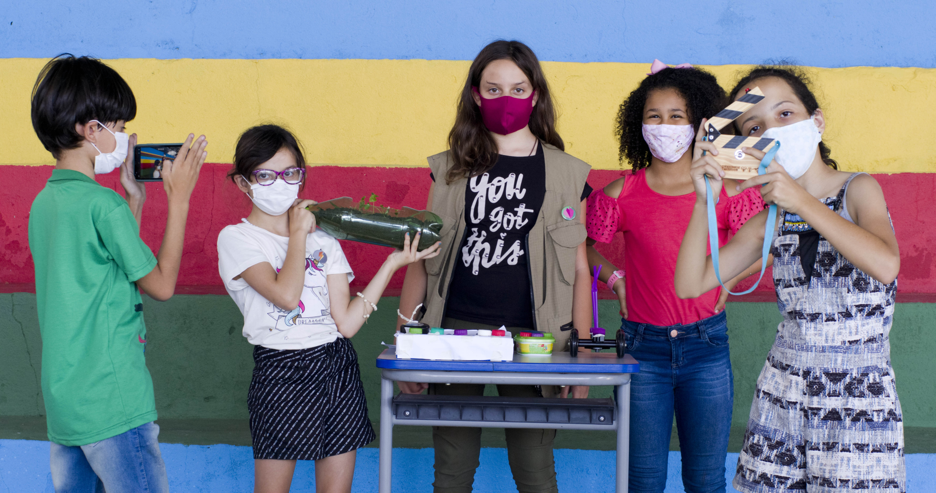 Cinco crianças com diversos experimentos e aparelhos em mãos olham para quem as fotografa.