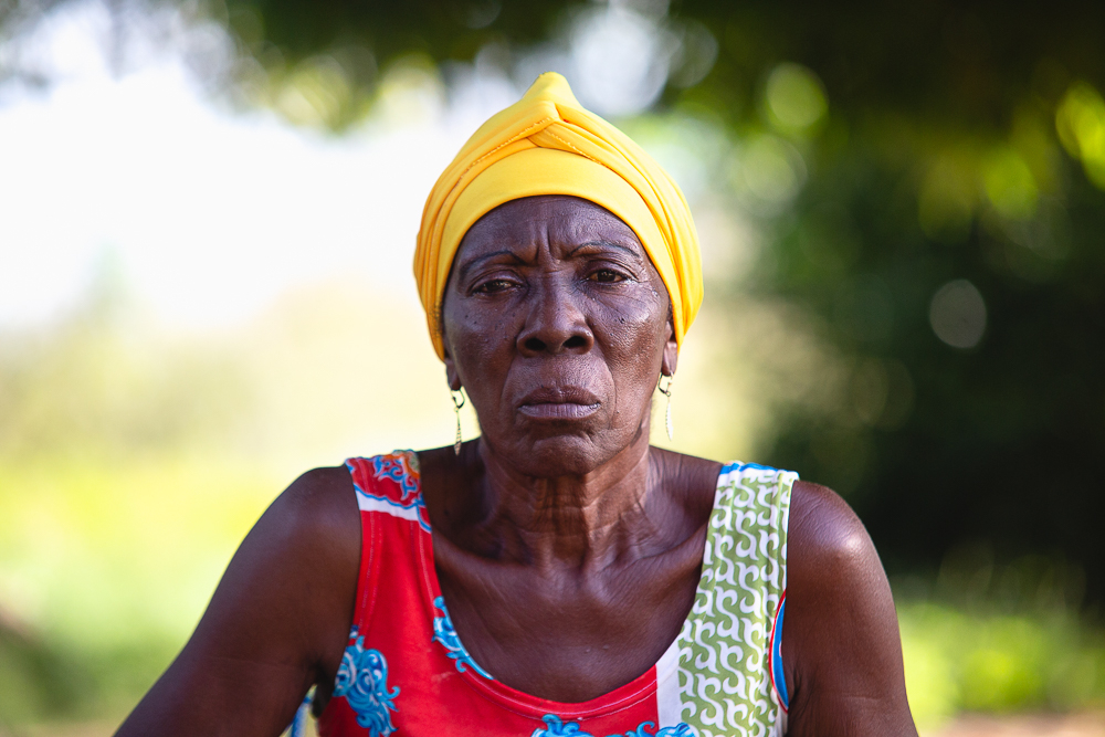 Uma mulher com olhar sério e expressão forte usa um turbante amarelo, olhando fixamente para quem a fotografou. Ao fundo, árvores.