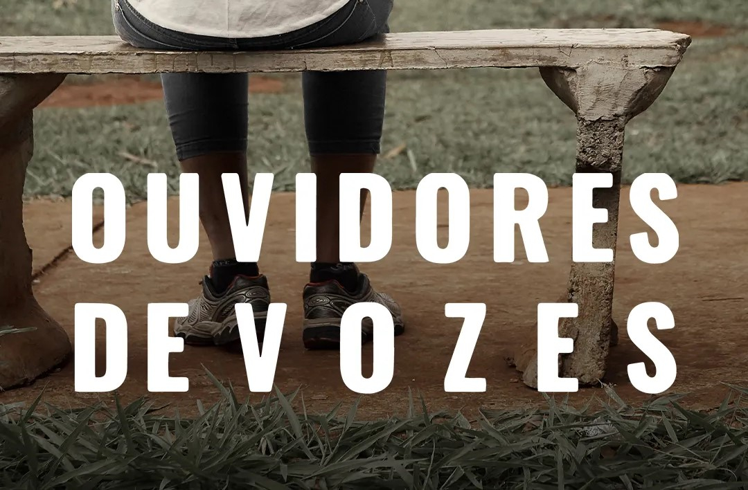 O título do filme "Ouvidores de vozes" sobre uma foto das pernas de uma pessoa que está sentada em um banco de madeira (a visão é de trás dessa pessoa).